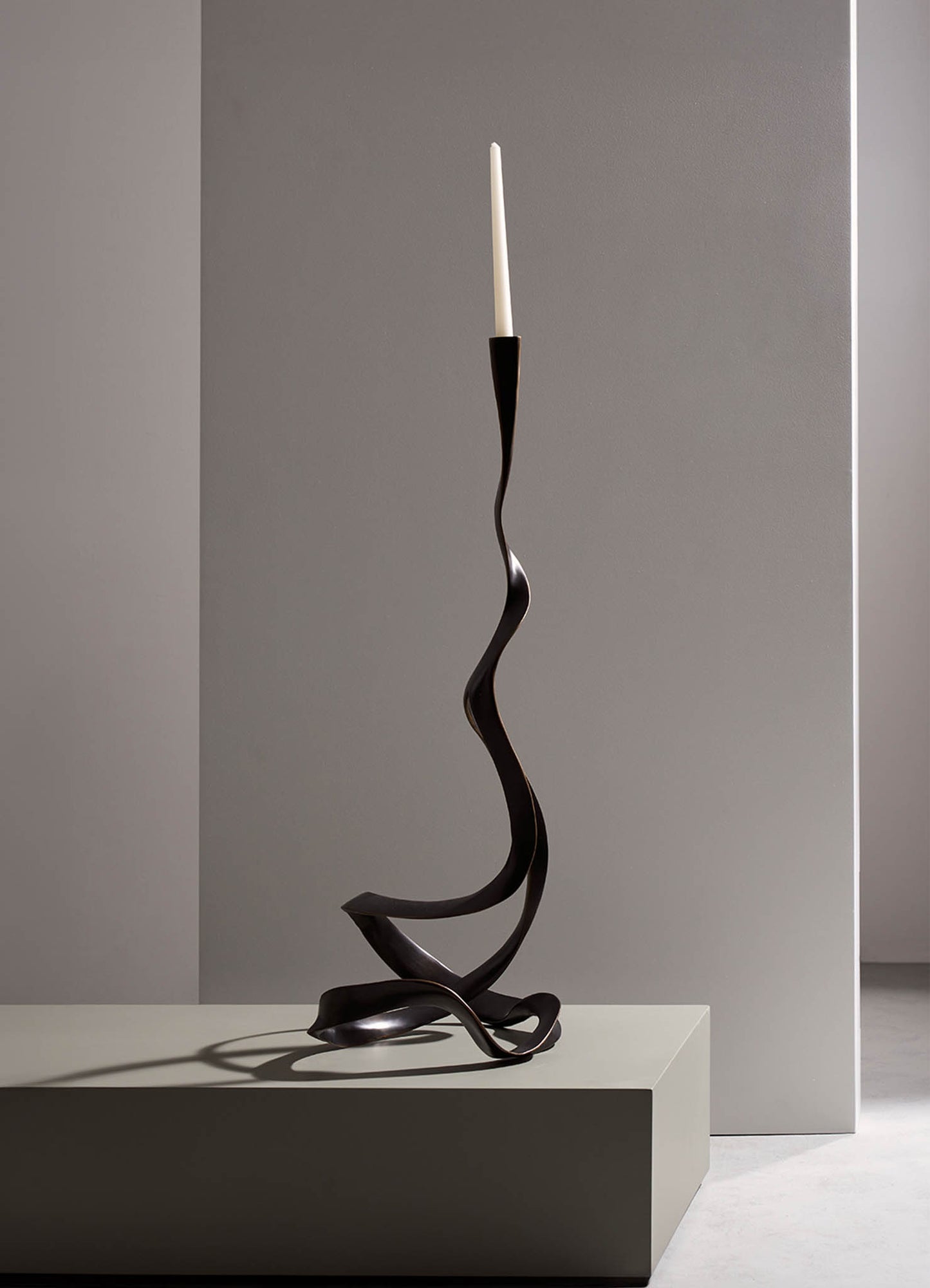 A sculptural candle holder with a black, twisted metal design and a white taper candle on top, set against a neutral gray background. The piece has an abstract, modern style with flowing, organic lines.
