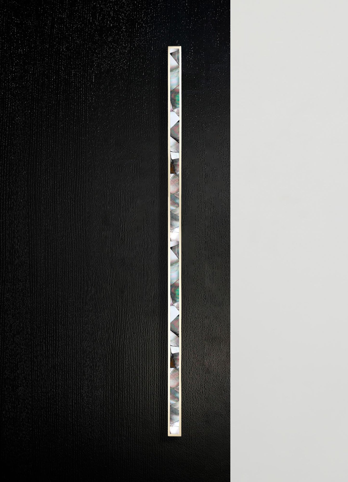 A vertical strip of iridescent, multicolored mother-of-pearl tiles set into a sleek, thin brass frame, mounted on a black textured surface with a contrasting smooth white background.
