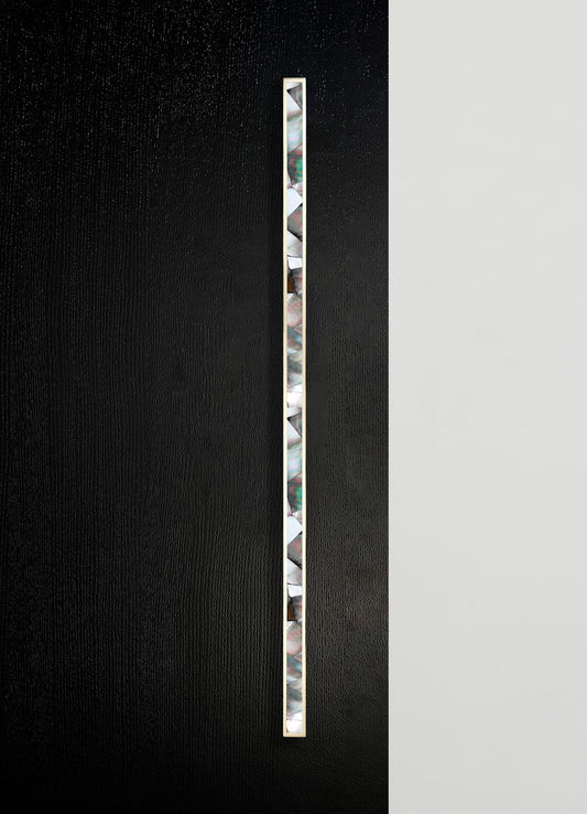 A vertical strip of iridescent, multicolored mother-of-pearl tiles set into a sleek, thin brass frame, mounted on a black textured surface with a contrasting smooth white background.