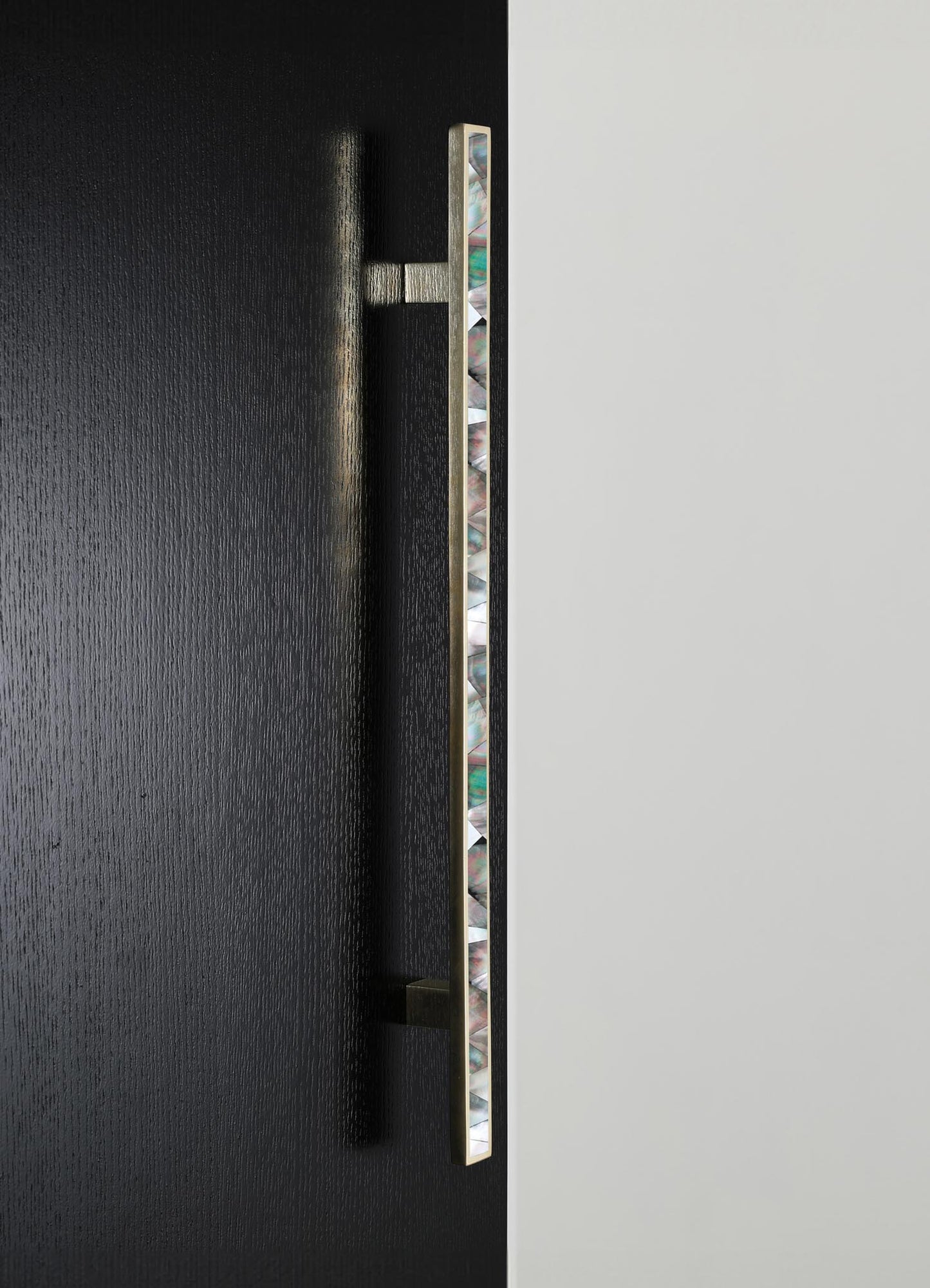 A modern door handle with an elongated brass design featuring an inlay of iridescent shell or mother-of-pearl motifs, mounted on a black wooden door with a textured finish.