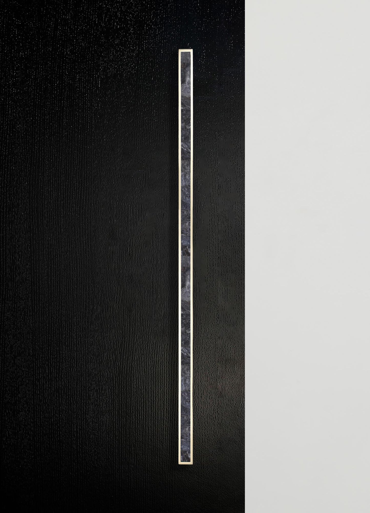 A vertical strip of dark marble with subtle veining is set on a textured black surface, bordered by a thin metallic edge. The design style is contemporary with a minimalist and sleek aesthetic.