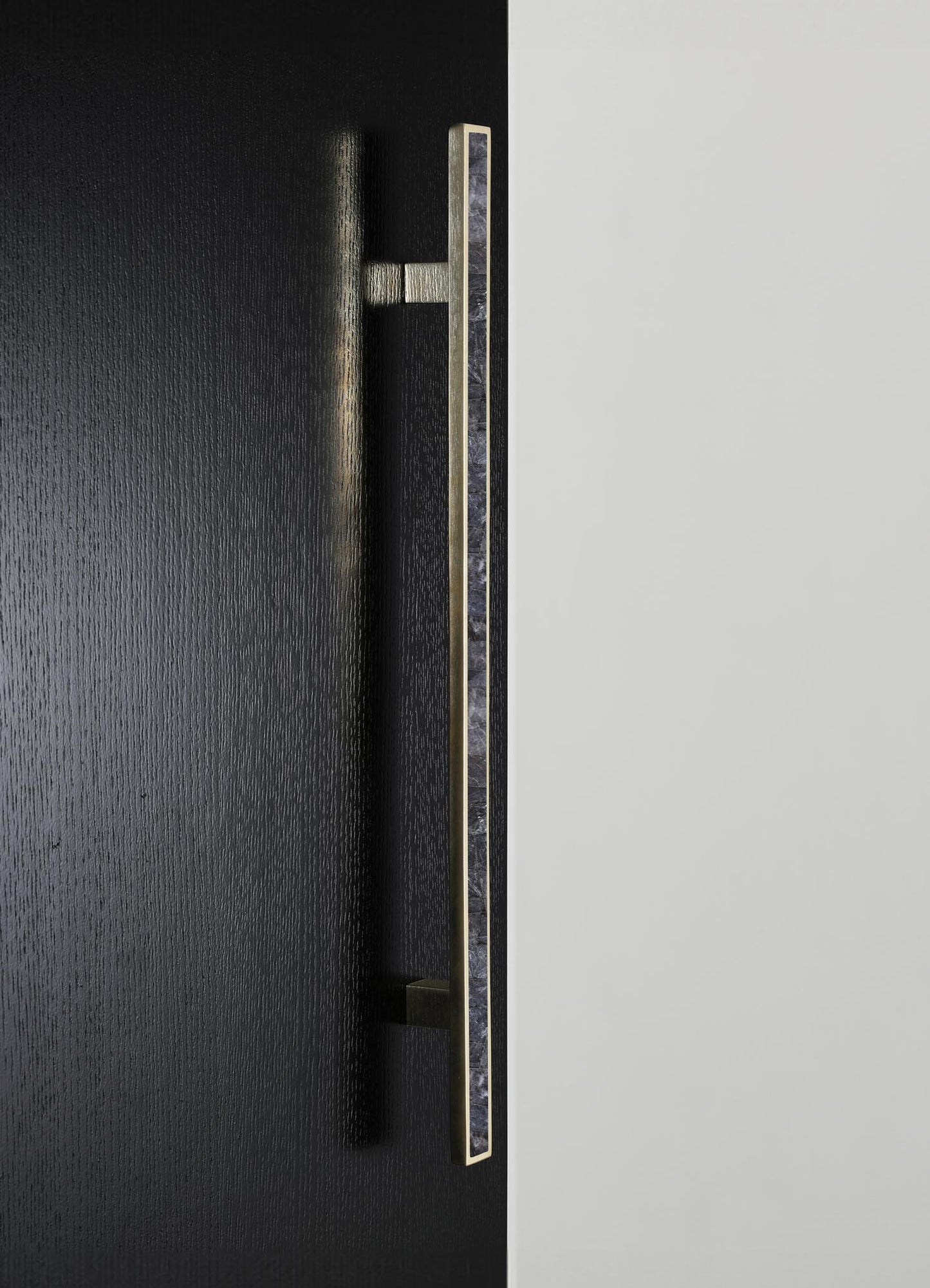 A close-up of a modern door handle made of brass or gold-toned metal with a textured black wall and a smooth light-colored wall background. The handle features a sleek, minimalist design with a rectangular profile and a geometric pattern on the visible side.