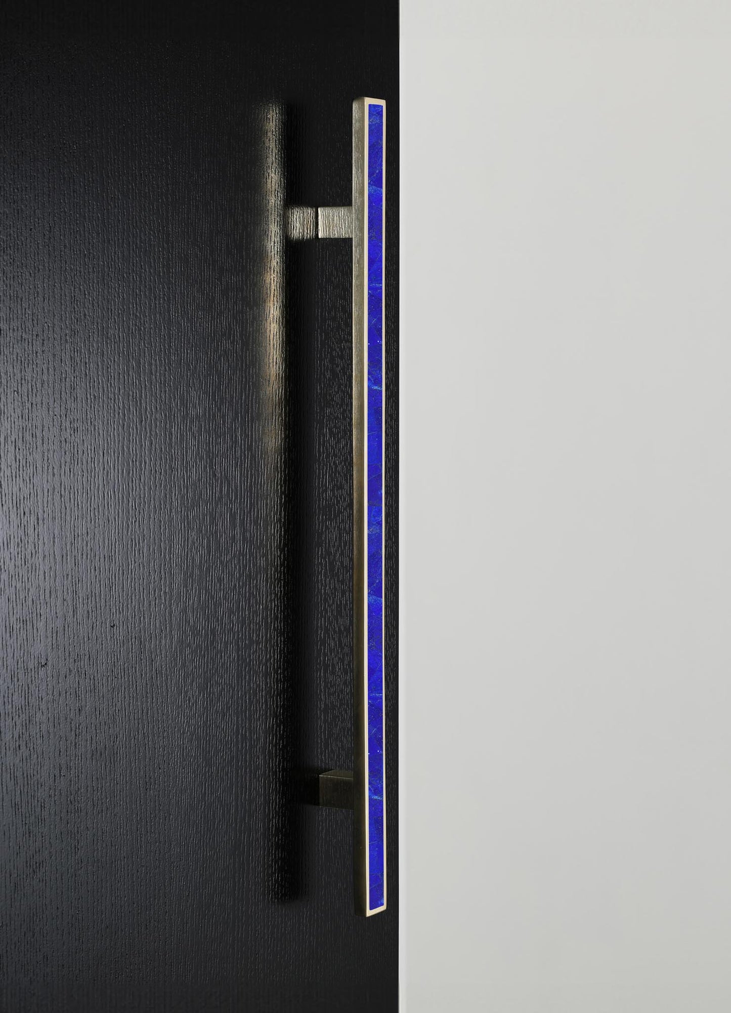 A vertical metal door handle with a sleek, modern design featuring a blue inlay detail, mounted on a black textured door. The handle has a minimalist aesthetic, emphasizing contemporary style and clean lines.