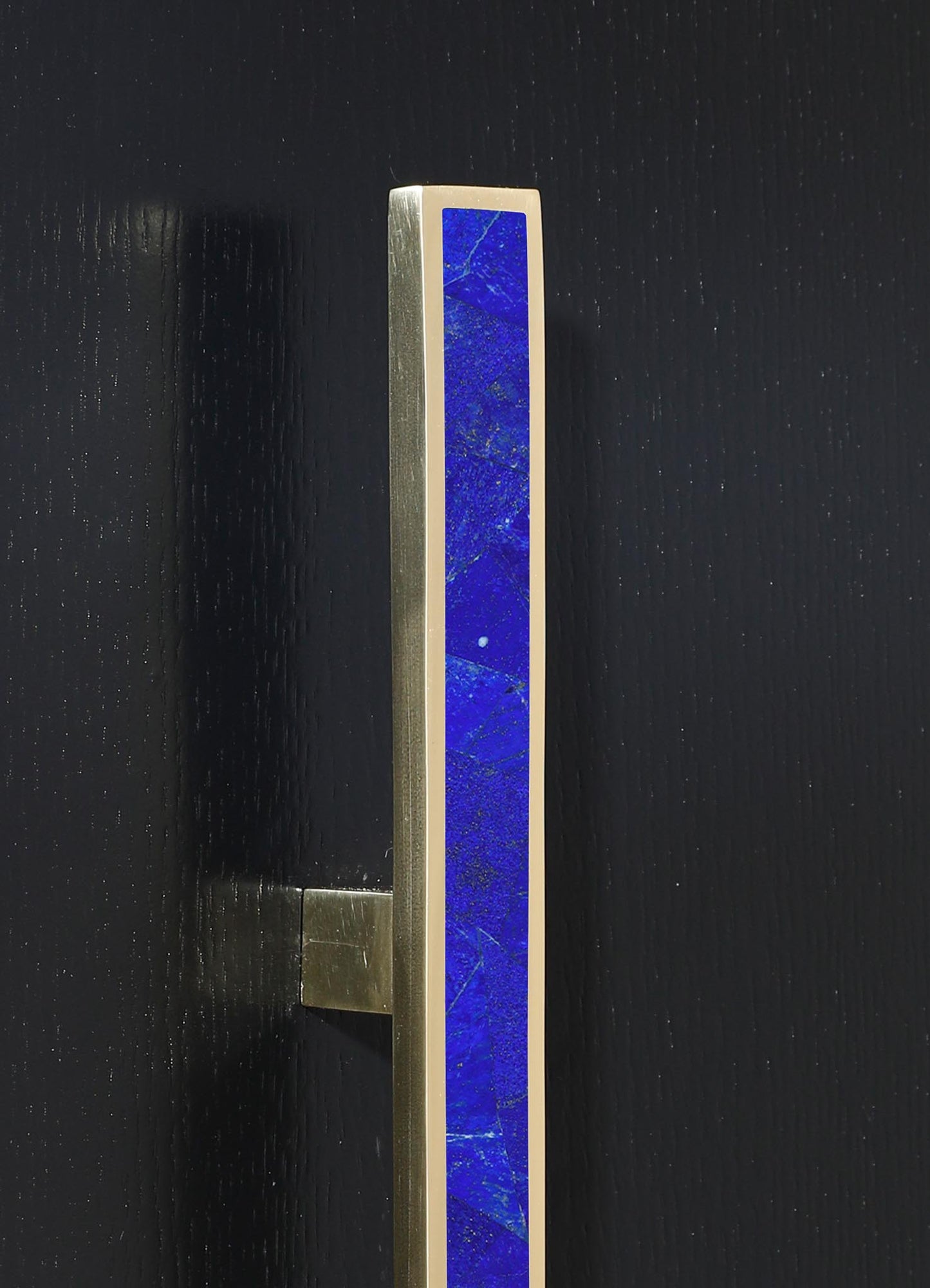 A modern door handle with a rectangular brass base, featuring a central inlay of vibrant blue lapis lazuli stone with natural gold and white veining, mounted on a black textured surface.