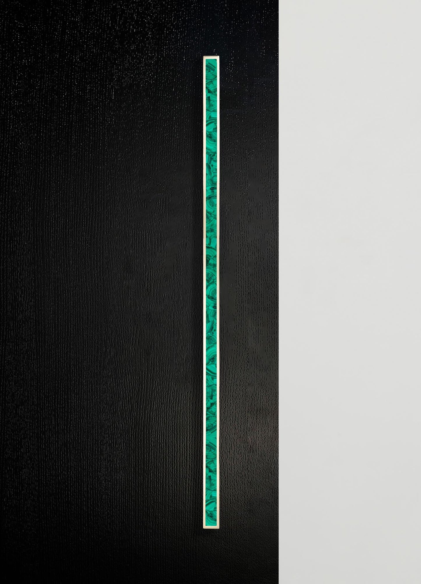 A vertical strip of layered malachite with shades of green and swirling patterns, set within a thin metallic frame on a textured black background. The design has a contemporary and minimalist style with a focus on natural stone motifs.