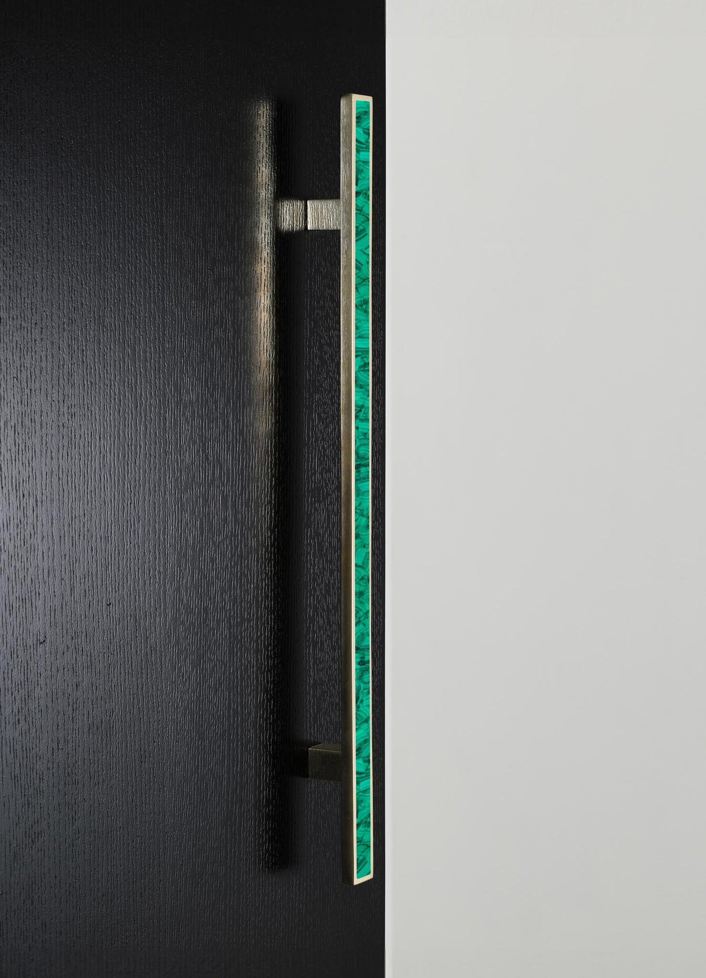 A modern door handle with a rectangular brass body featuring a vertical strip of green malachite inlay. The handle is mounted on a black textured surface, contrasting with a smooth light-colored background.