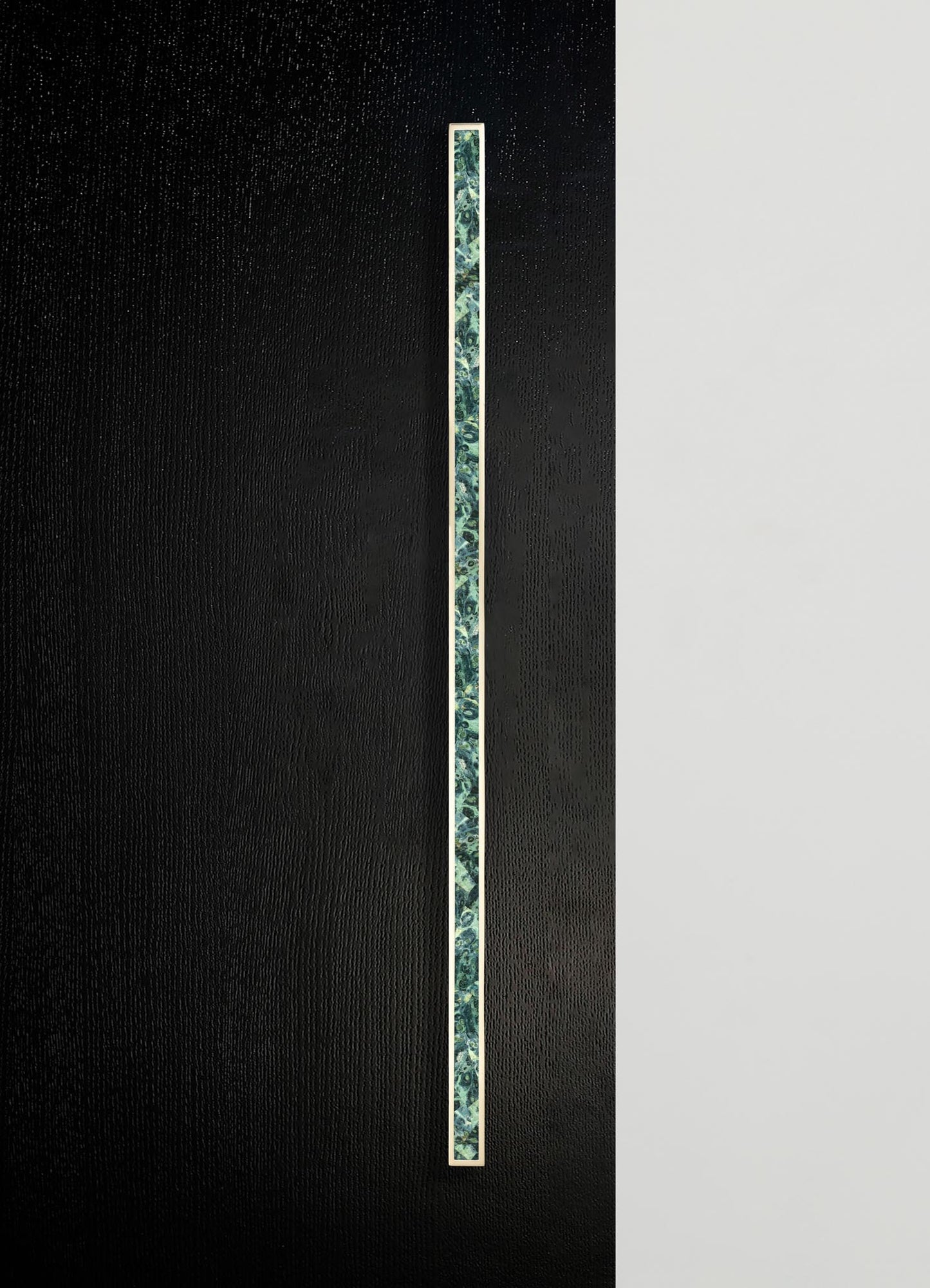 A vertical strip of patterned ceramic tile with blue and green floral motifs, set against a textured black background and a plain white surface. The tile has a narrow metallic border framing the design.