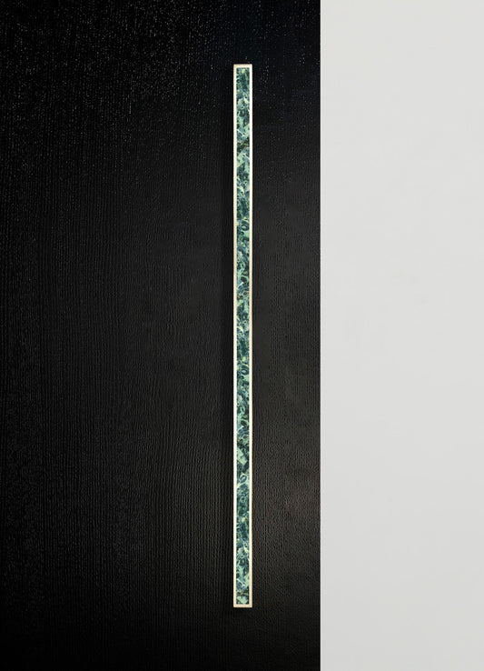 A vertical strip of patterned ceramic tile with blue and green floral motifs, set against a textured black background and a plain white surface. The tile has a narrow metallic border framing the design.