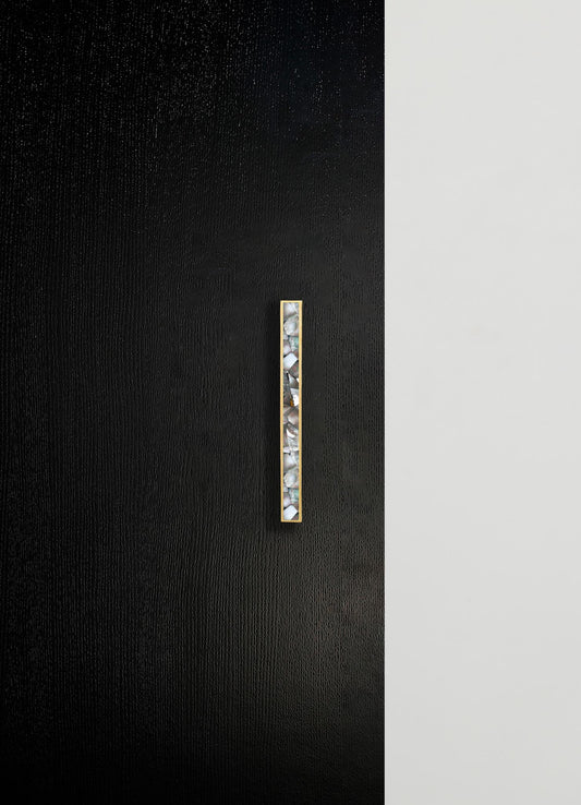 Minimalist wall decor featuring a narrow vertical inlay with a textured, iridescent mosaic pattern set in a slim metallic frame, contrasted against a black textured wall and a smooth white section.