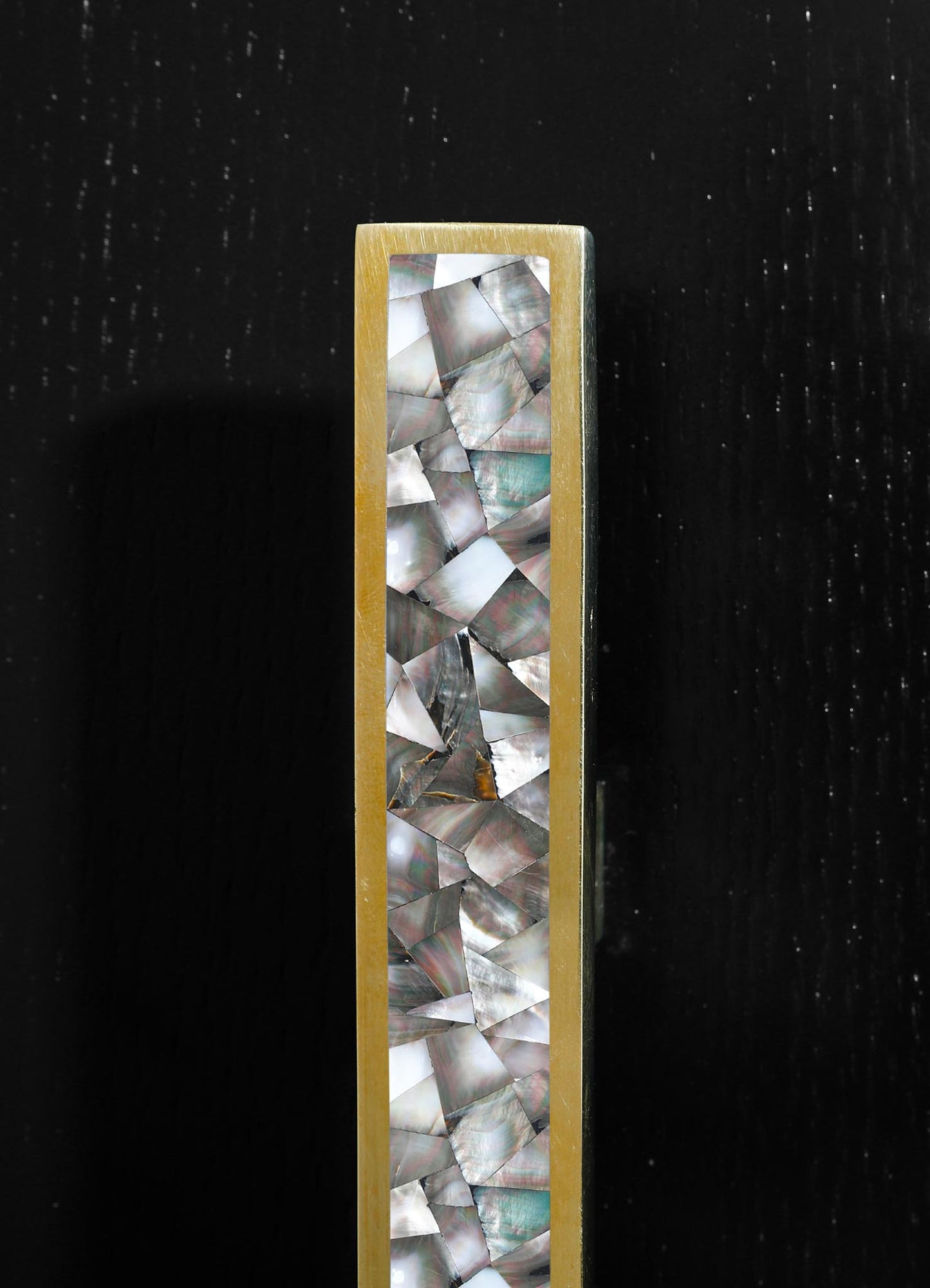 A tall, narrow decorative piece with a brass frame featuring shell or mother-of-pearl inlays arranged in a geometric pattern, set against a black background.
