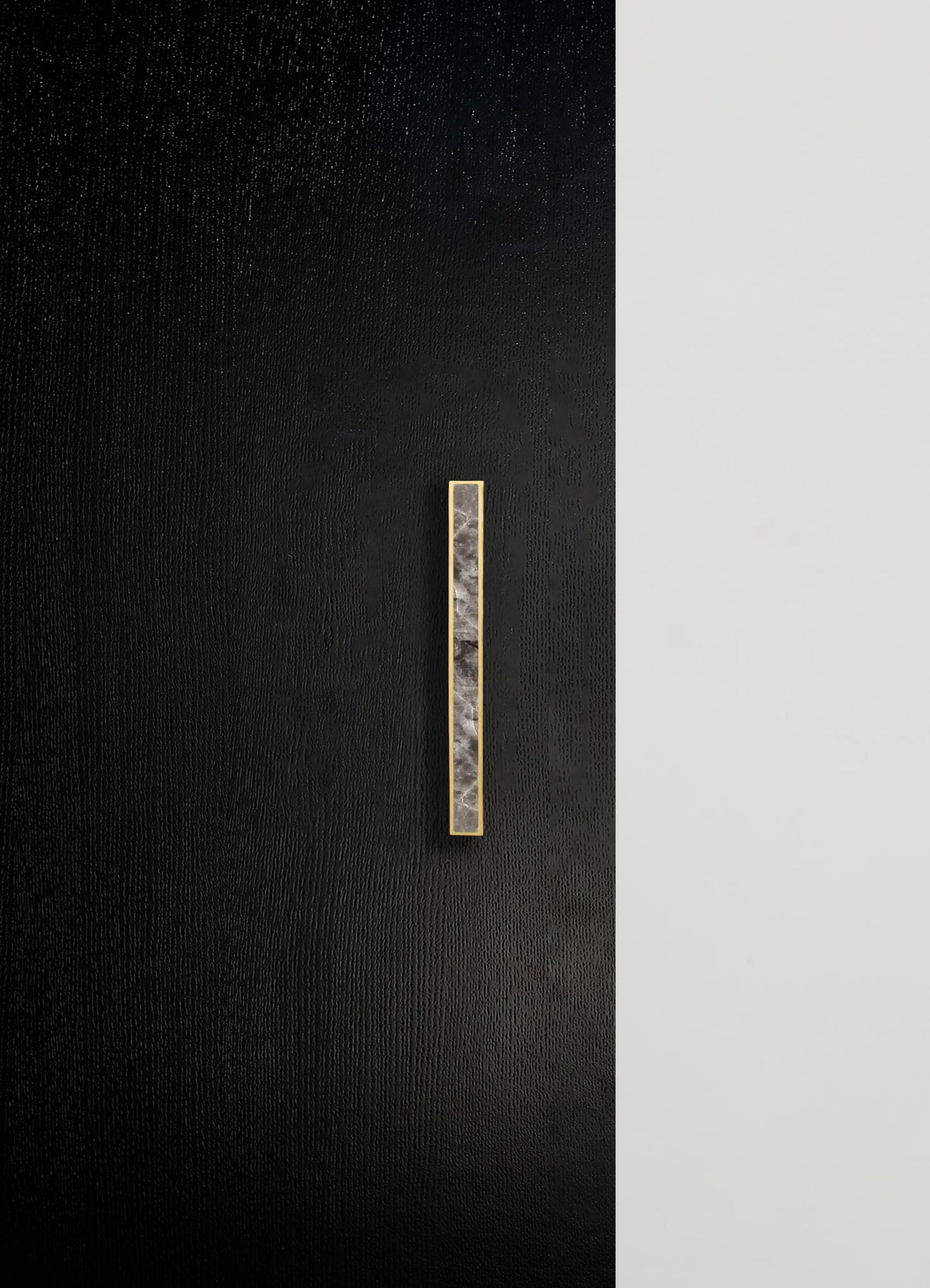 A minimalist wall design featuring a black textured surface on the left and a smooth white surface on the right, with a vertical marble and brass wall handle or accent strip positioned on the black side.