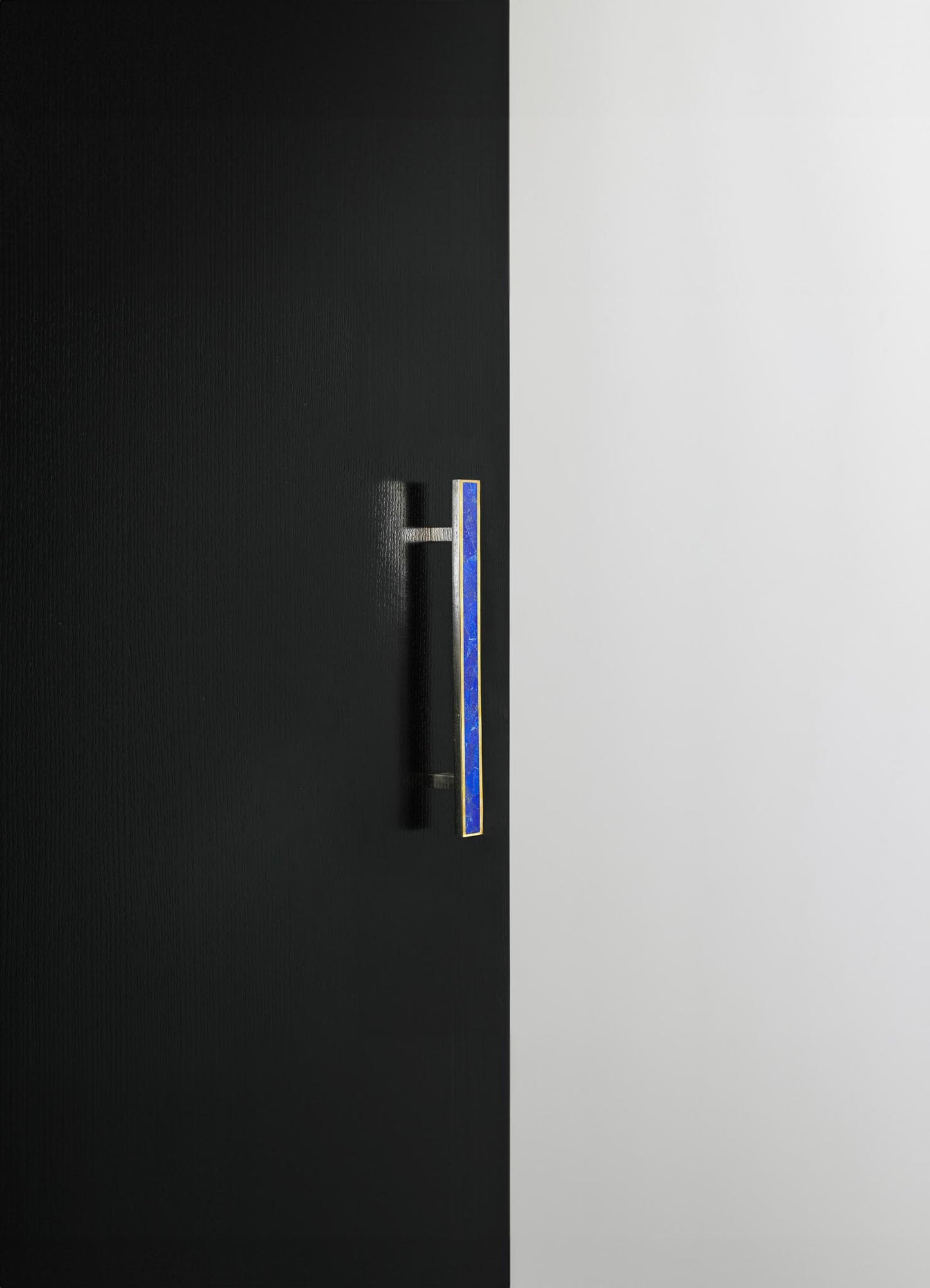 A door with a textured black surface featuring a slim vertical handle with blue and gold accents, set against a smooth white wall. The design has a modern and minimalist style.