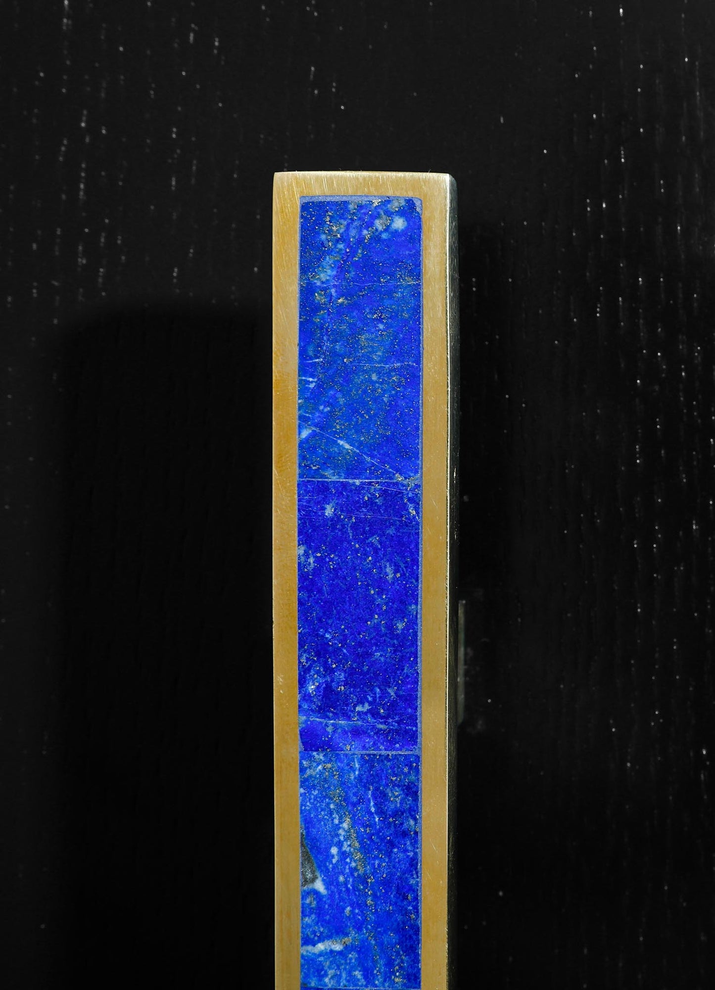 A tall brass candle holder with a rectangular shape featuring a vivid blue lapis lazuli inlay, set against a black background. The design emphasizes a contemporary style with a luxurious and polished finish.