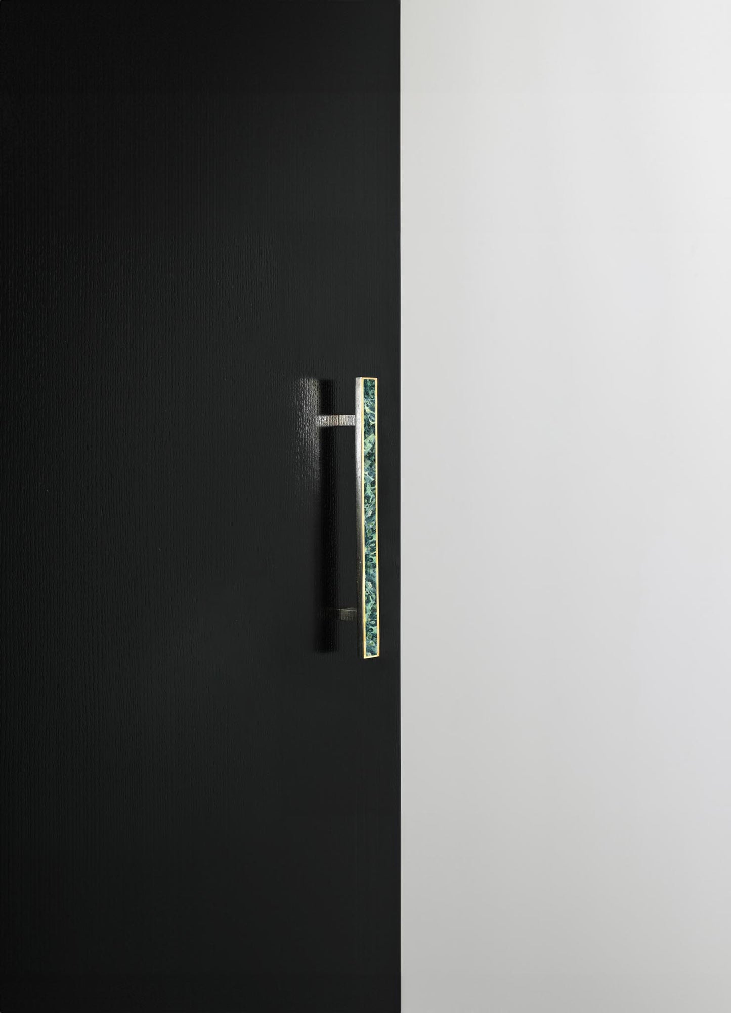 A modern black door with a textured surface and a vertical brass handle featuring a marbled green and blue pattern, set against a smooth white wall. The design emphasizes minimalism and contemporary style.