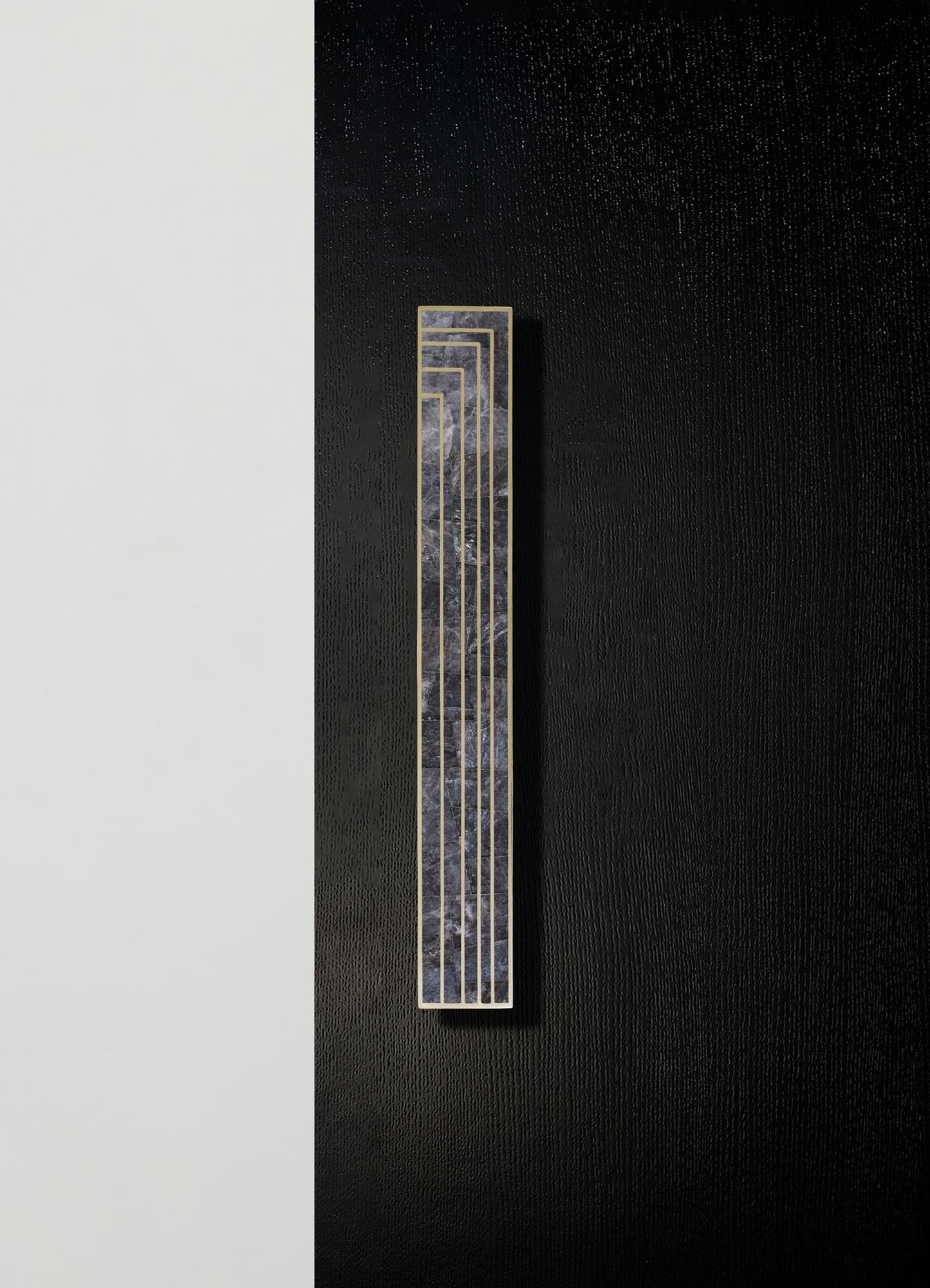 A vertical wall-mounted sculpture combining an abstract marble pattern with a geometric gold frame, set against a textured black wall with a contrasting white section.