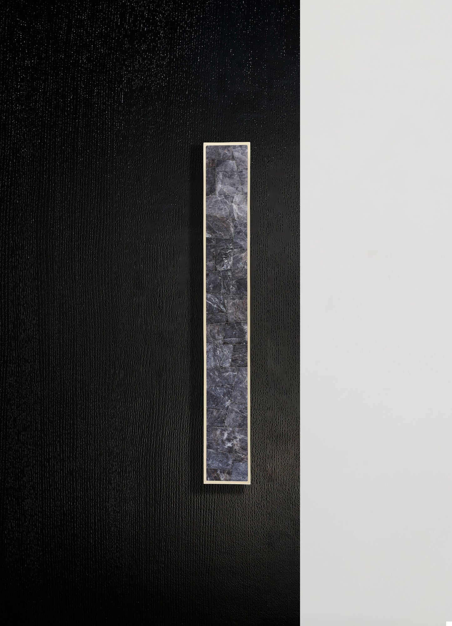 A narrow, vertical panel featuring a mosaic of dark gray stone tiles with a textured surface, framed by a thin metallic border, mounted on a black and white textured wall.