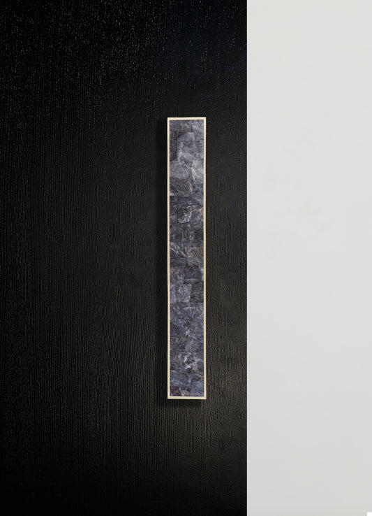 A narrow, vertical panel featuring a mosaic of dark gray stone tiles with a textured surface, framed by a thin metallic border, mounted on a black and white textured wall.
