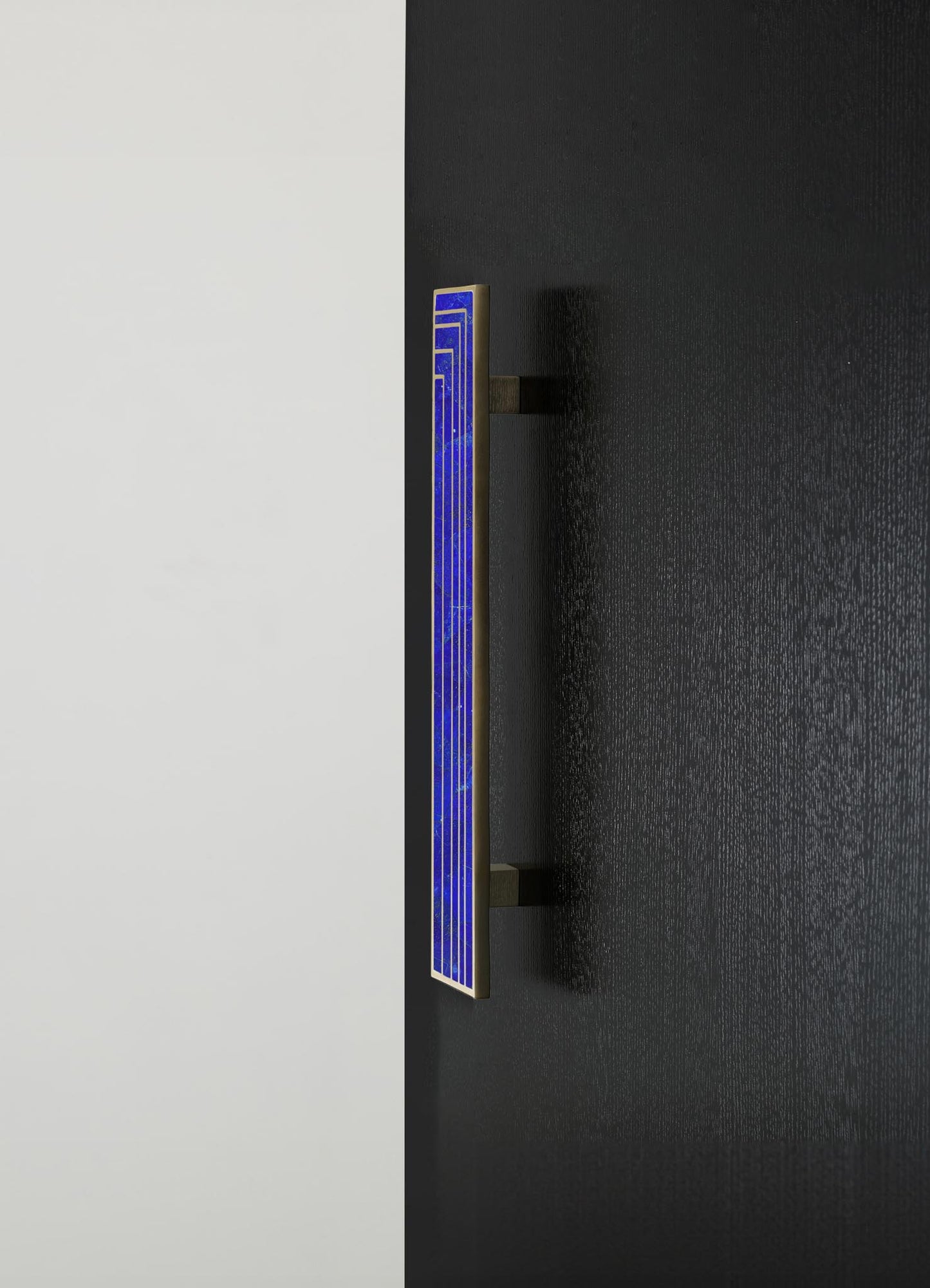 A modern wall sconce featuring a rectangular metal frame with a blue glass panel and geometric pattern, mounted on a textured black wall with a contrasting white background.