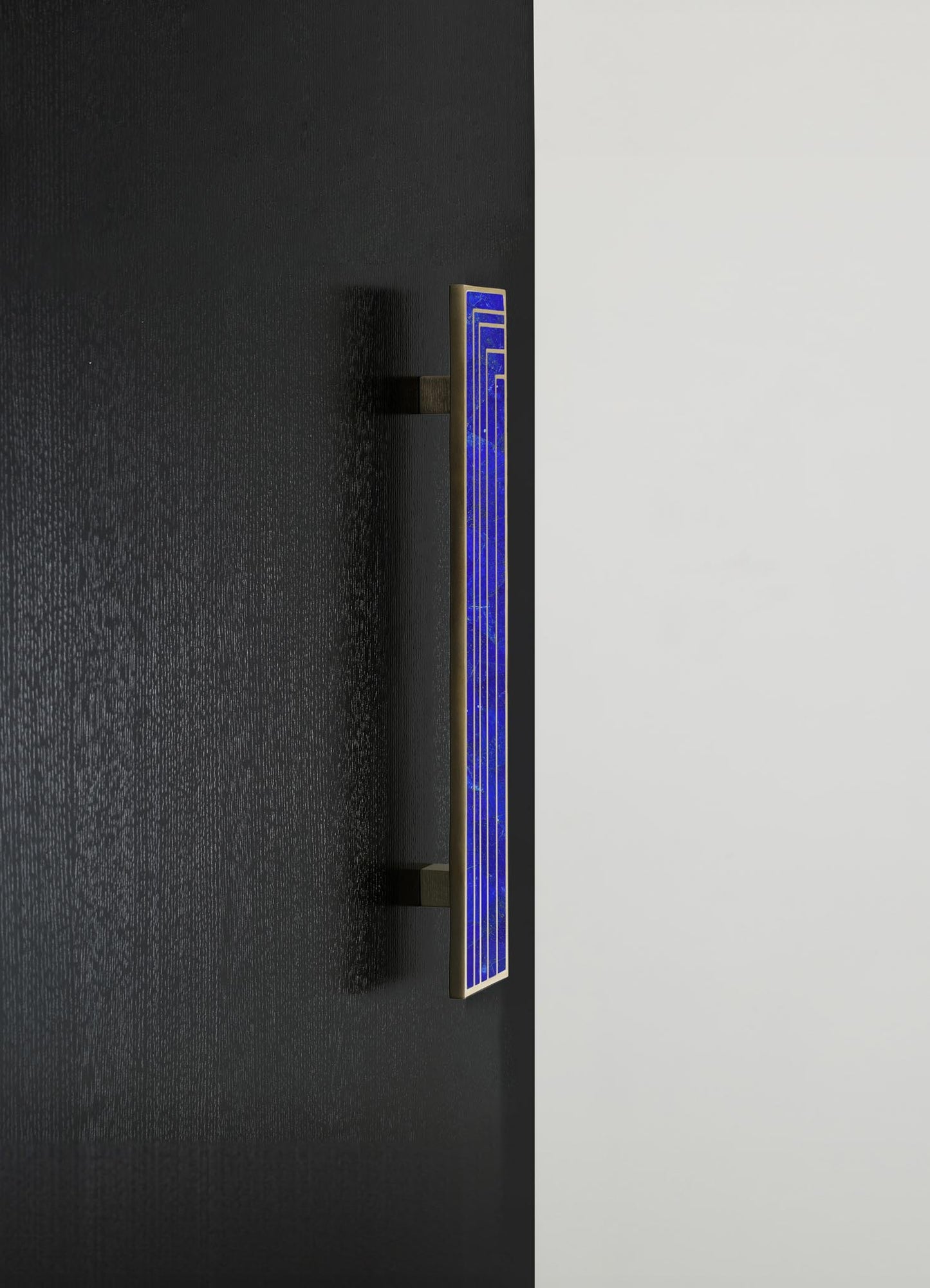 A modern wall-mounted LED light fixture with a vertical rectangular shape, featuring a vibrant blue glass panel and a metallic brass frame. The fixture is installed on a black wall next to a white wall, emphasizing contemporary minimalistic design and sleek materials.