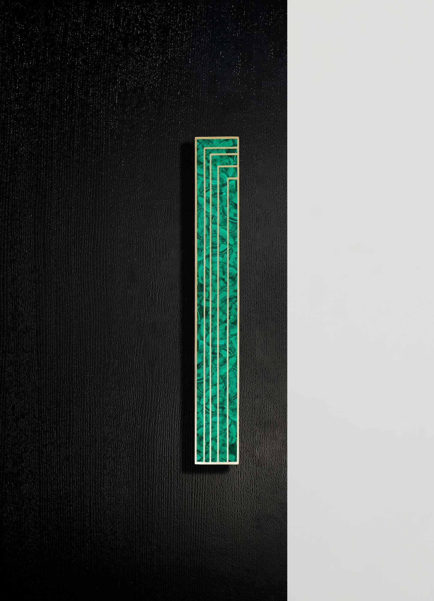 A vertical decorative panel featuring a geometric pattern in shades of green, framed with a light-colored border, mounted on a textured black wall with a contrasting plain white section.
