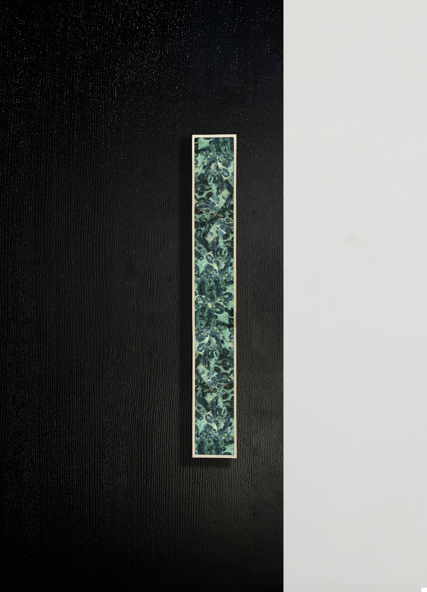 A rectangular decorative wall panel with a floral and paisley pattern in shades of green, teal, and black, framed with a thin white border. The background features a textured black surface on the left and a smooth white surface on the right.