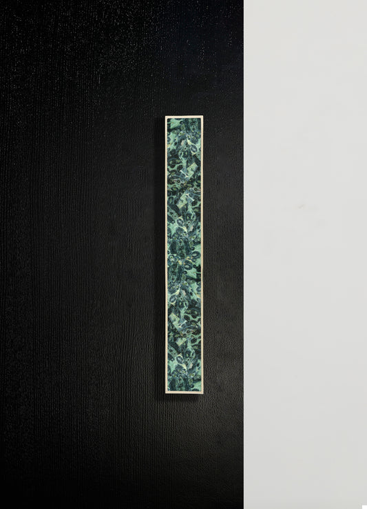 A rectangular decorative wall panel with a floral and paisley pattern in shades of green, teal, and black, framed with a thin white border. The background features a textured black surface on the left and a smooth white surface on the right.