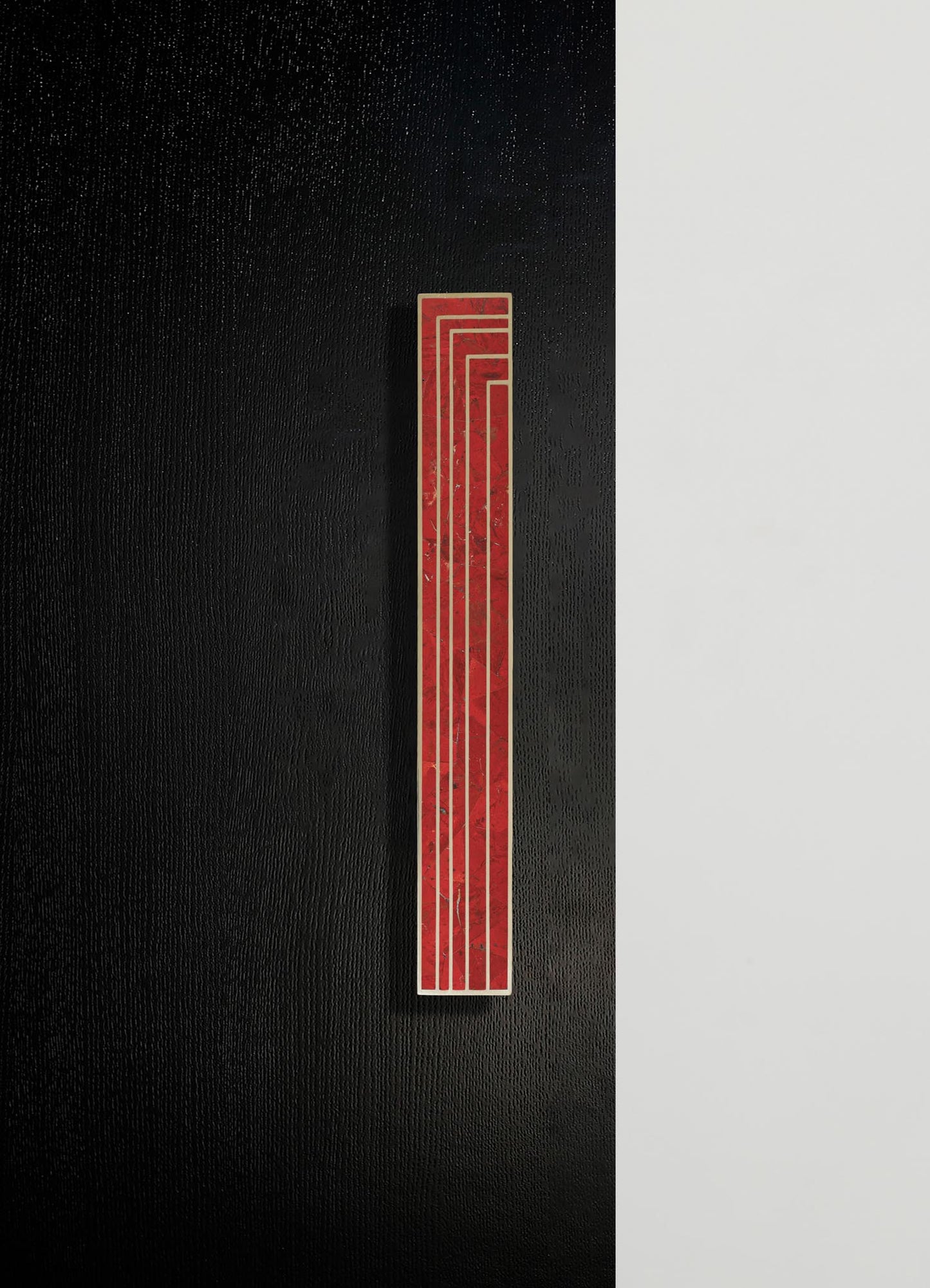 A wall-mounted abstract art piece featuring a narrow rectangular design with red marble texture and gold lines, set against a black textured wall divided by a white panel in a contemporary interior.