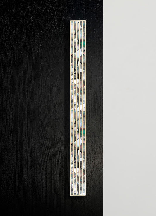 A tall, narrow decorative object featuring a white frame with an inner mosaic of iridescent, geometrically arranged shell or glass pieces, set against a split black and white background.