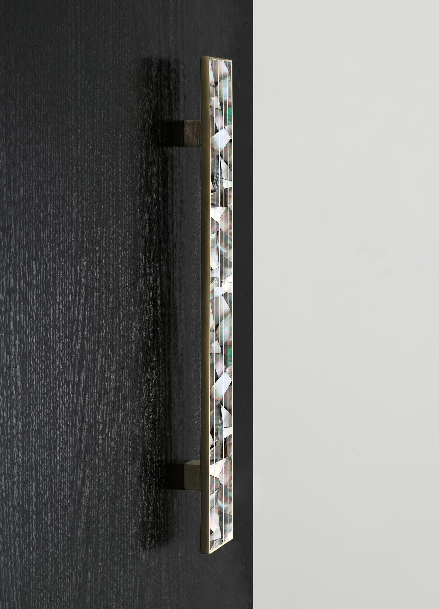 A vertical wall-mounted light fixture with a slim brass frame and geometric mosaic pattern of iridescent glass tiles, set against contrasting black and white walls. The design has a contemporary style with a focus on modern minimalist decor.