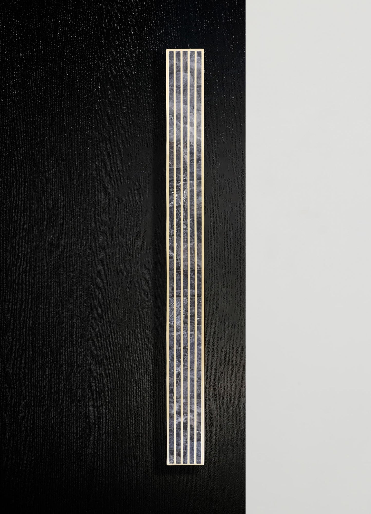 A tall, rectangular wall sconce with a brass frame and a textured, translucent glass or acrylic panel, mounted on a black and white wall. The design is modern and minimalist, emphasizing metallic accents and geometric form.