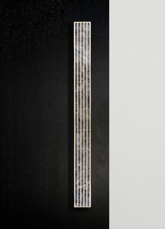 A tall, rectangular wall sconce with a brass frame and a textured, translucent glass or acrylic panel, mounted on a black and white wall. The design is modern and minimalist, emphasizing metallic accents and geometric form.
