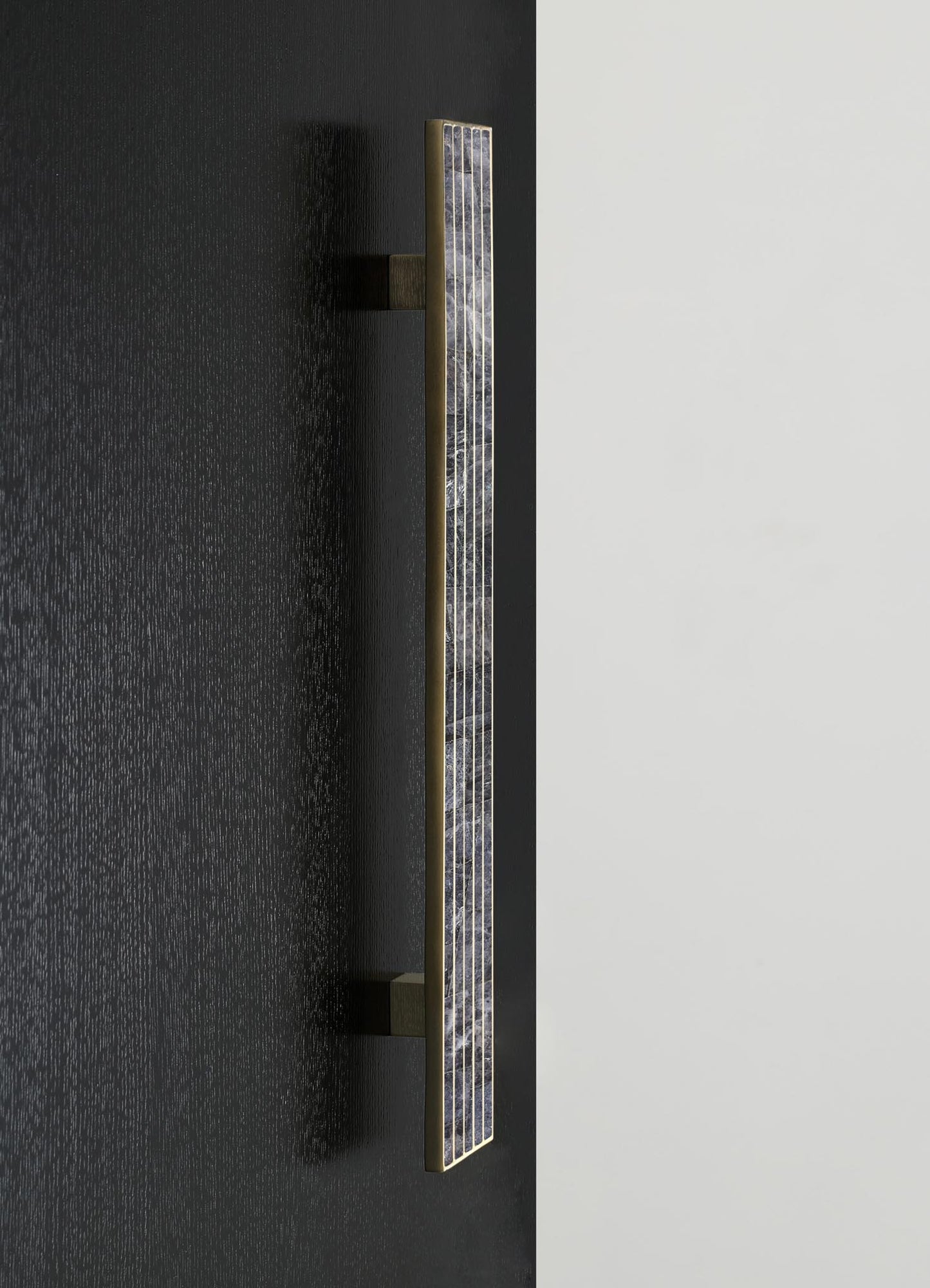 A modern wall-mounted light fixture featuring a slim, elongated design with a brass finish and geometric patterned glass or acrylic panels, set against a dark wall with contrasting light-colored background.