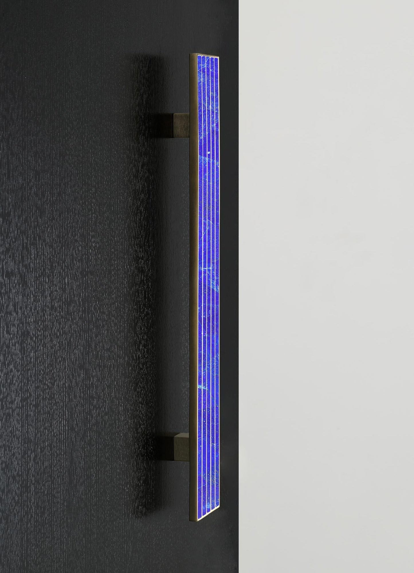 A modern wall-mounted LED light fixture with a slim, rectangular shape featuring a blue illuminated panel and a brass or gold-toned frame, set against a black and white wall background.