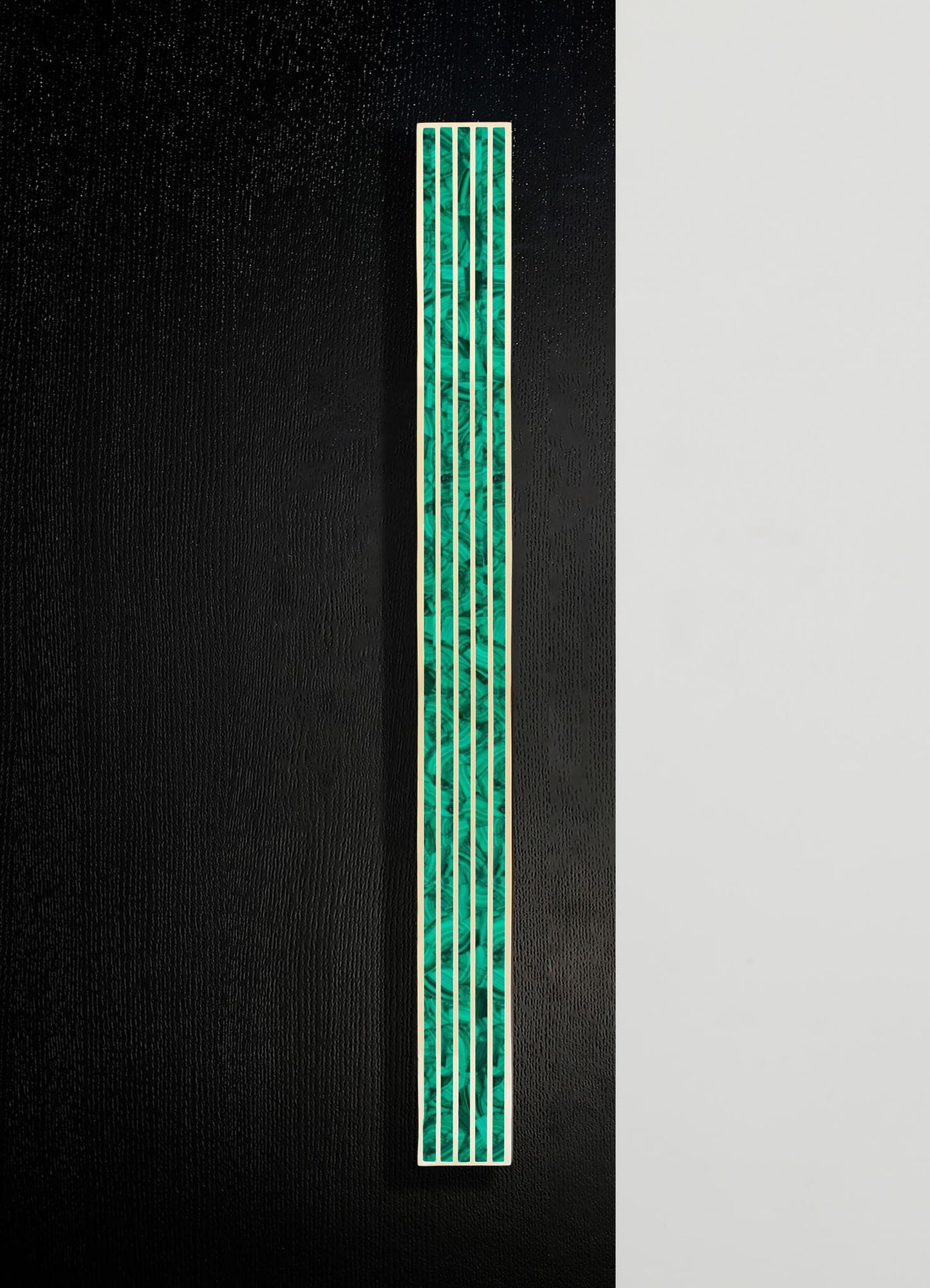 A tall decorative panel featuring an abstract mosaic with green and teal tones, framed in white and mounted on a black textured wall. The design has a contemporary style with a sleek and artistic appearance.