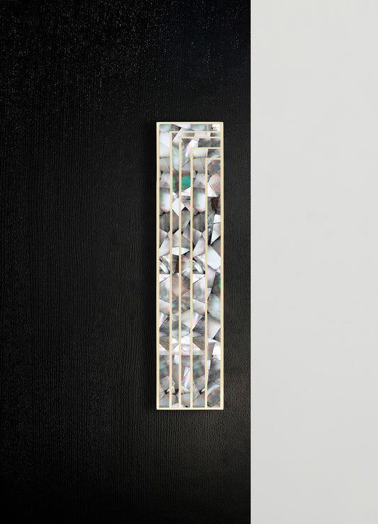 A decorative wall panel with a geometric lattice design made of metal with a gold finish, featuring an inlaid abalone shell pattern with iridescent, multicolored tones. The panel is installed on a wall with contrasting black and white textured surfaces.