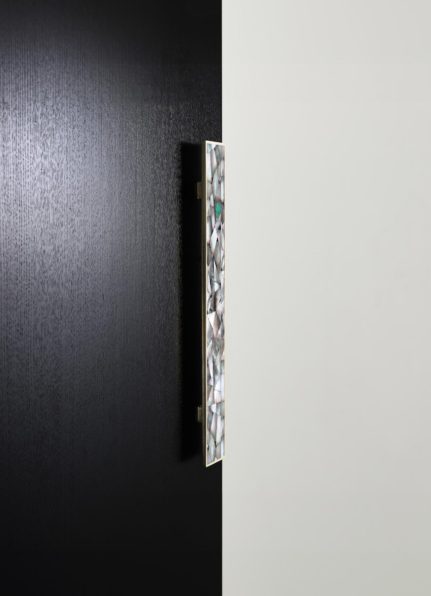 A vertical decorative wall panel with an iridescent, shell-like pattern set in a metallic bezel, mounted on a black textured wall. The panel has a sleek, contemporary style with reflective motifs and metallic accents.