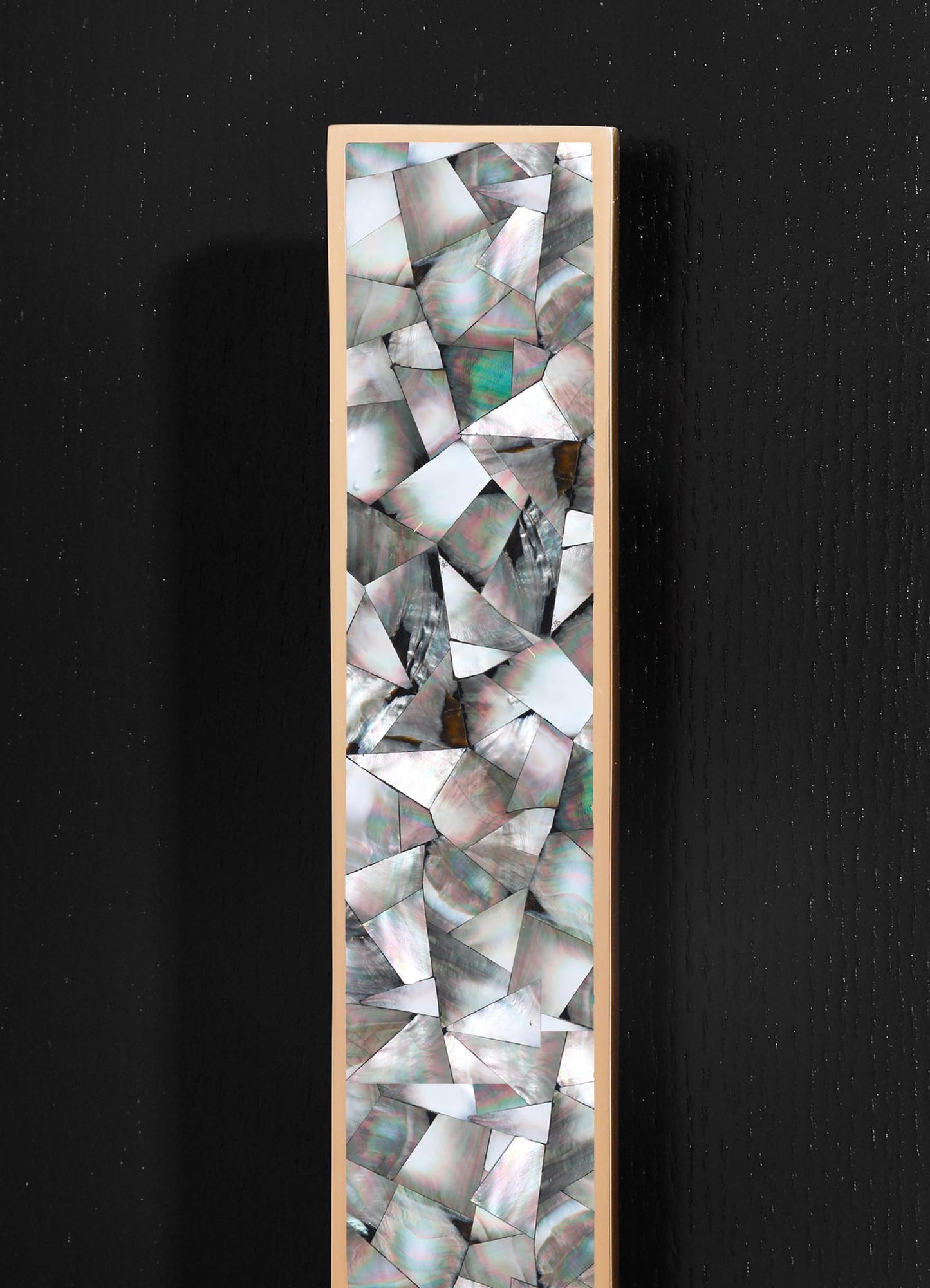 A tall, rectangular mirror with a slim, beige frame featuring a mosaic pattern of abalone shell pieces in iridescent shades of gray, green, and pink, set against a black textured wall.