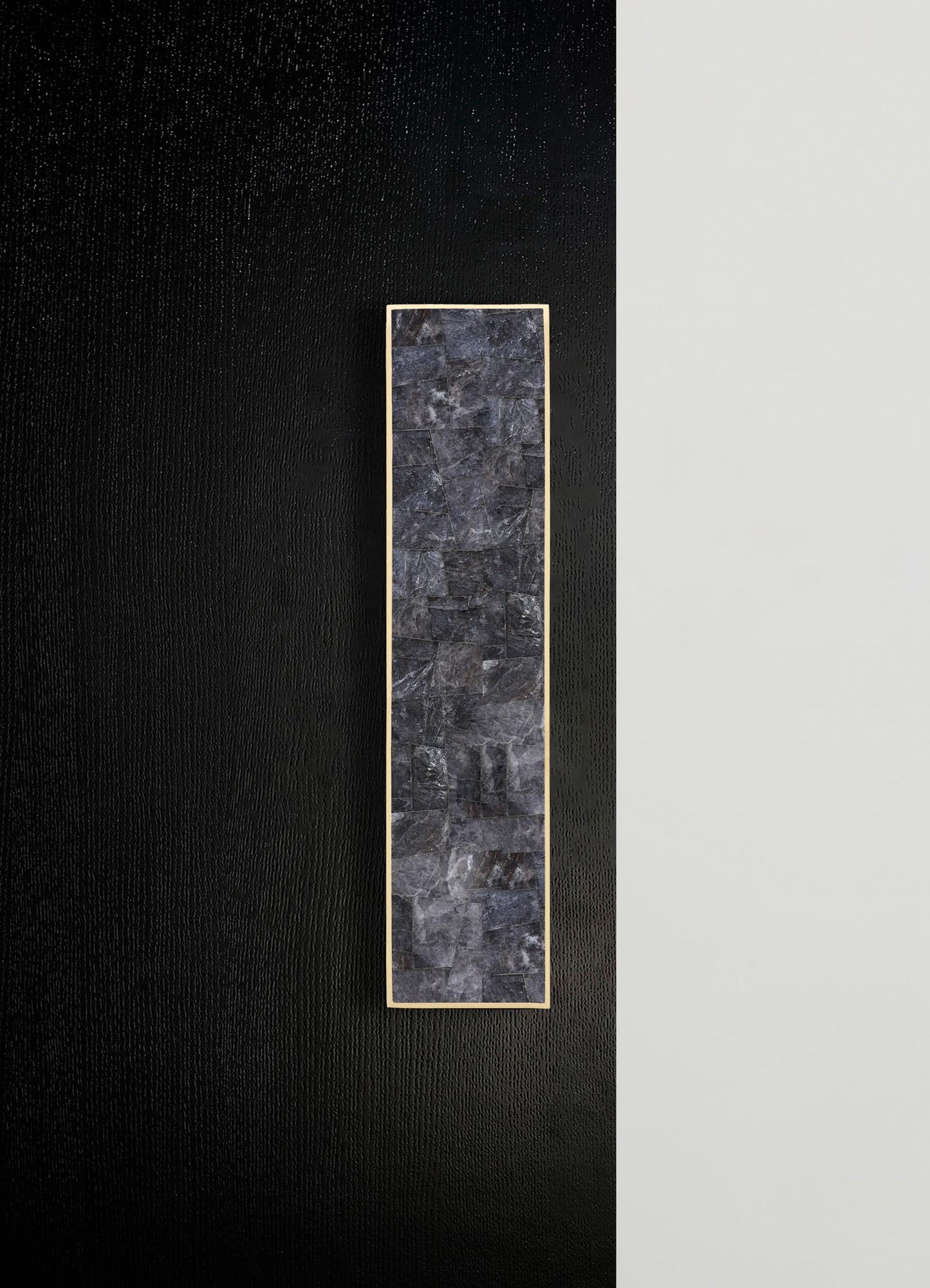 A vertical rectangular wall panel with a dark grey stone finish framed by a thin metallic border, mounted on a wall with contrasting black and white textured surfaces.