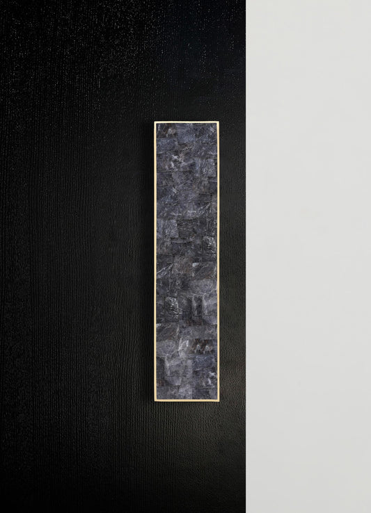A vertical rectangular wall panel with a dark grey stone finish framed by a thin metallic border, mounted on a wall with contrasting black and white textured surfaces.