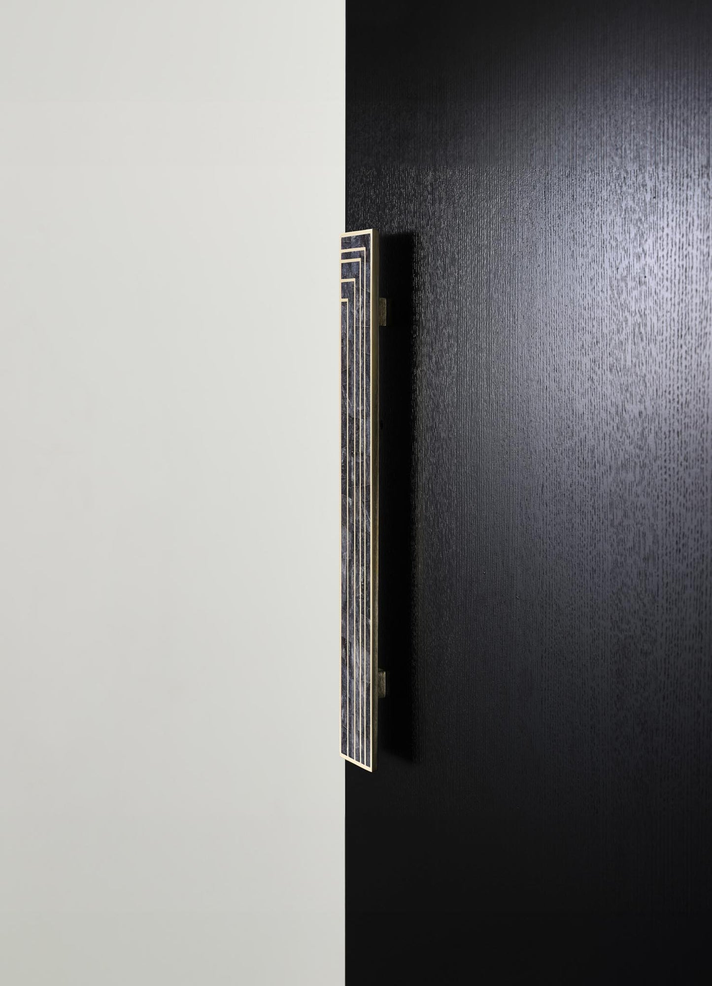 A modern wall sconce with a rectangular geometric design, featuring a brass or gold-tone metal frame and a dark textured shade, mounted on a black textured wall. The background includes a plain white wall, emphasizing the sleek, contemporary style of the fixture.
