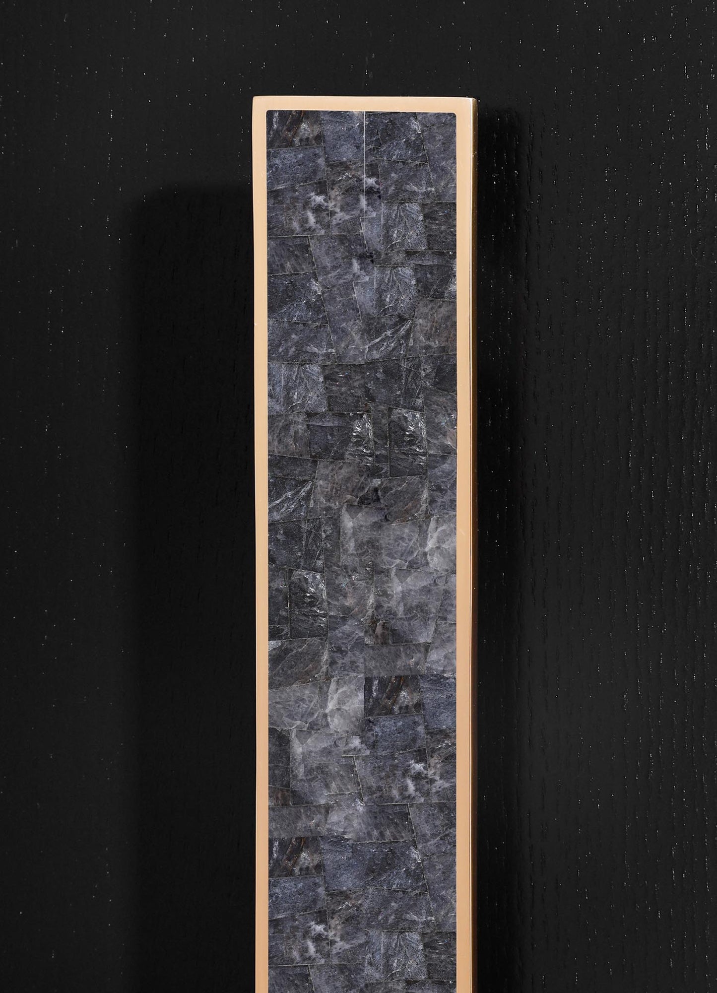 A tall, rectangular panel with a frame in a light beige or cream color surrounds a surface made of dark gray stone tiles, exhibiting a natural, textured pattern. The panel is mounted on a black wall with a subtle dotted texture.