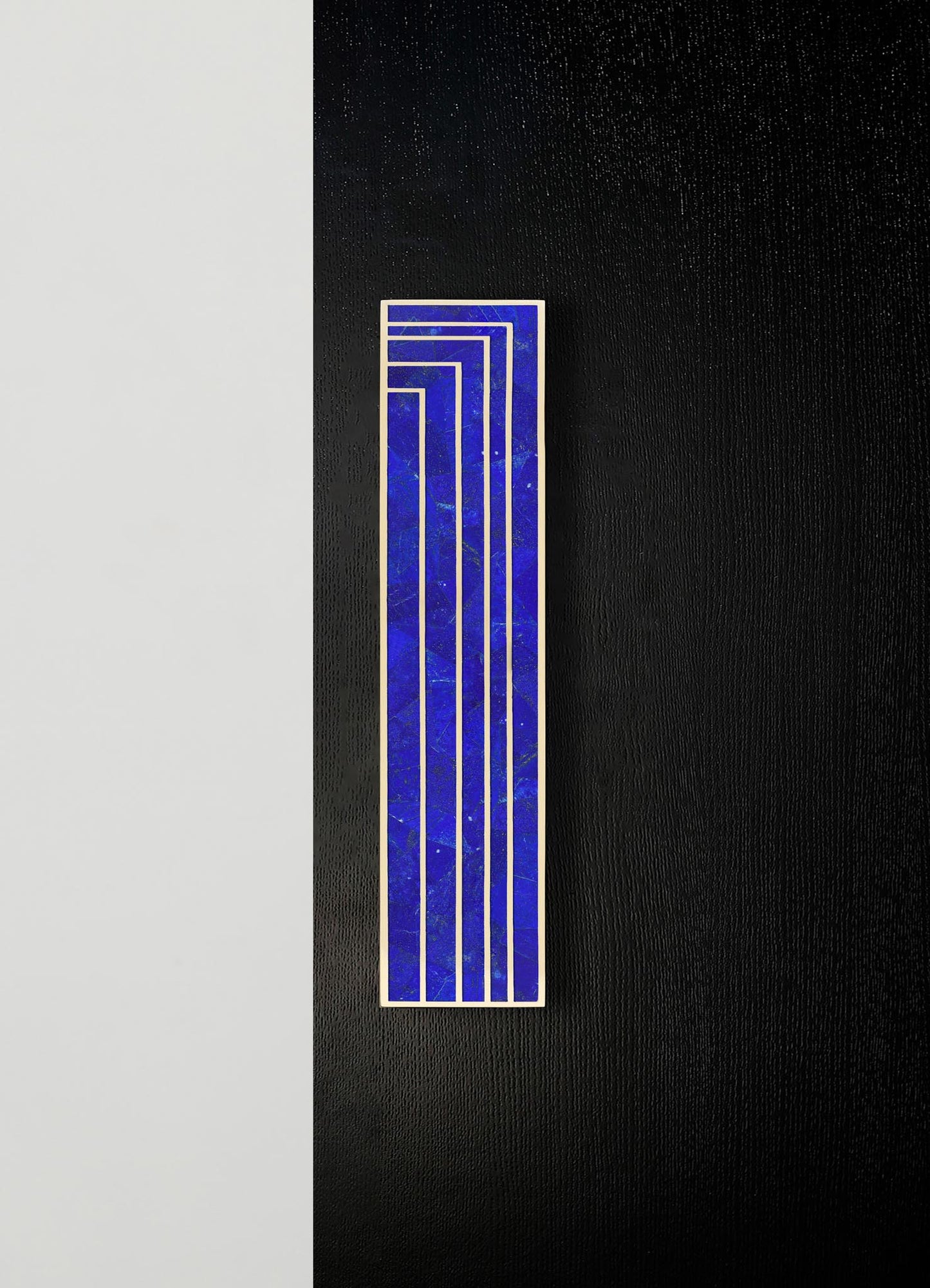 A decorative wall art piece featuring a rectangular panel with a geometric pattern of parallel lines in blue, set against a black textured wall with a contrasting light-colored surface nearby.