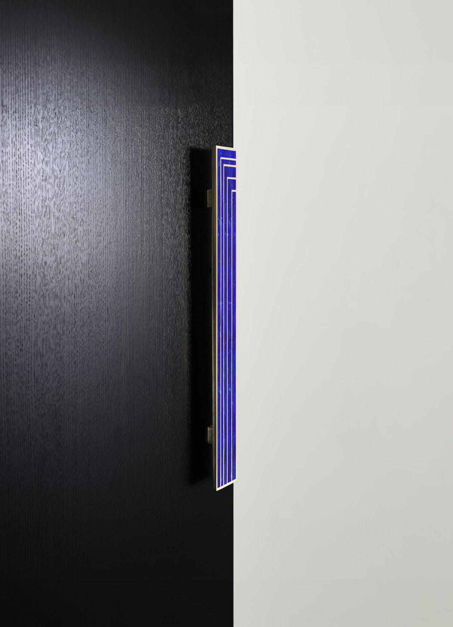 A modern wall art piece featuring a vertical rectangular panel with a blue geometric design, mounted on a black textured wall. The minimalistic style emphasizes clean lines and vibrant color contrast against the dark background.