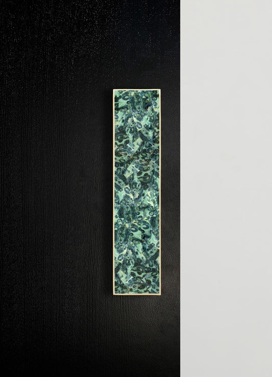A rectangular wall art piece featuring an abstract pattern with shades of blue, green, and black, framed with a slim light-colored border. The artwork is mounted against a black textured wall with a contrasting white panel on one side.