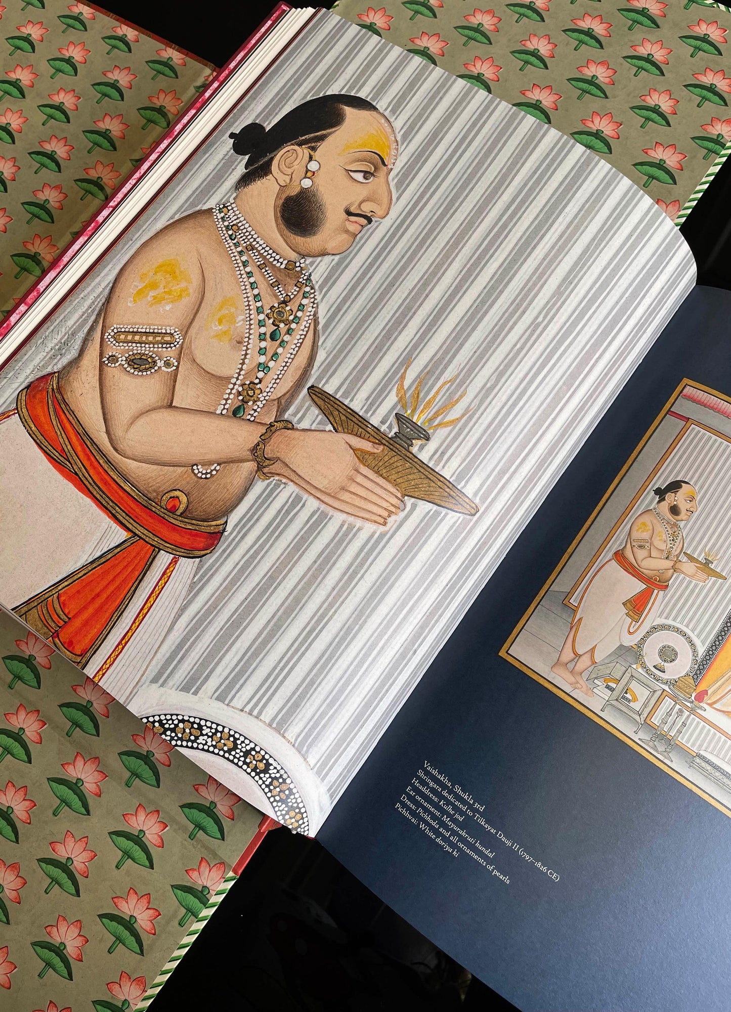 A hand-drawn illustration of a traditional Indian figure wearing jewelry and a dhoti, holding a lit oil lamp. The background features vertical gray and white stripes, and the figure is adorned with colorful clothing and ornate accessories.