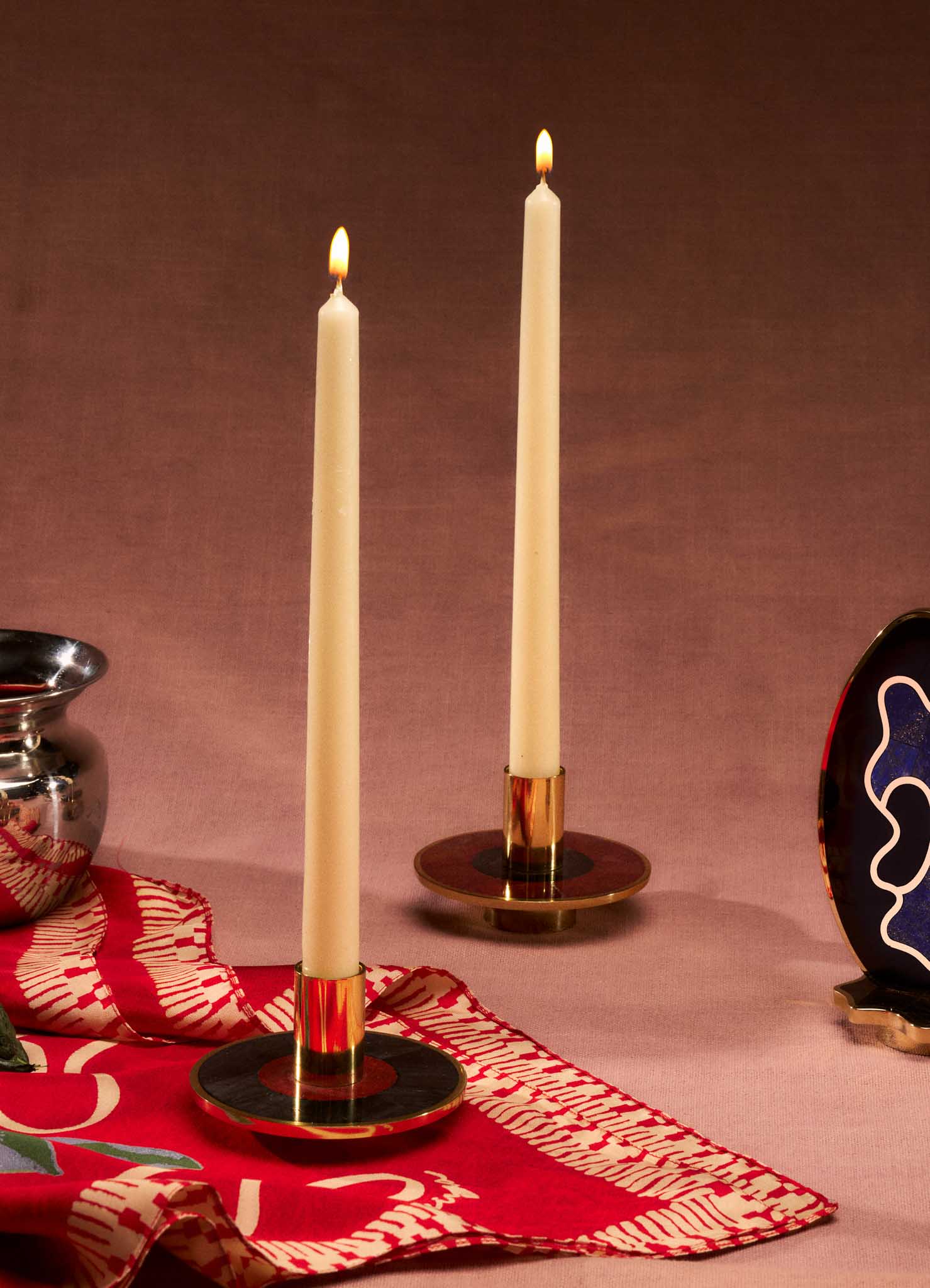 Two lit white taper candles in VIYA Alta Candle Stands sit on a red and white cloth, with a metallic vase, vibrant Betel leaves, and a colorful décor piece nearby, all set against a brown fabric backdrop for an auspicious life.