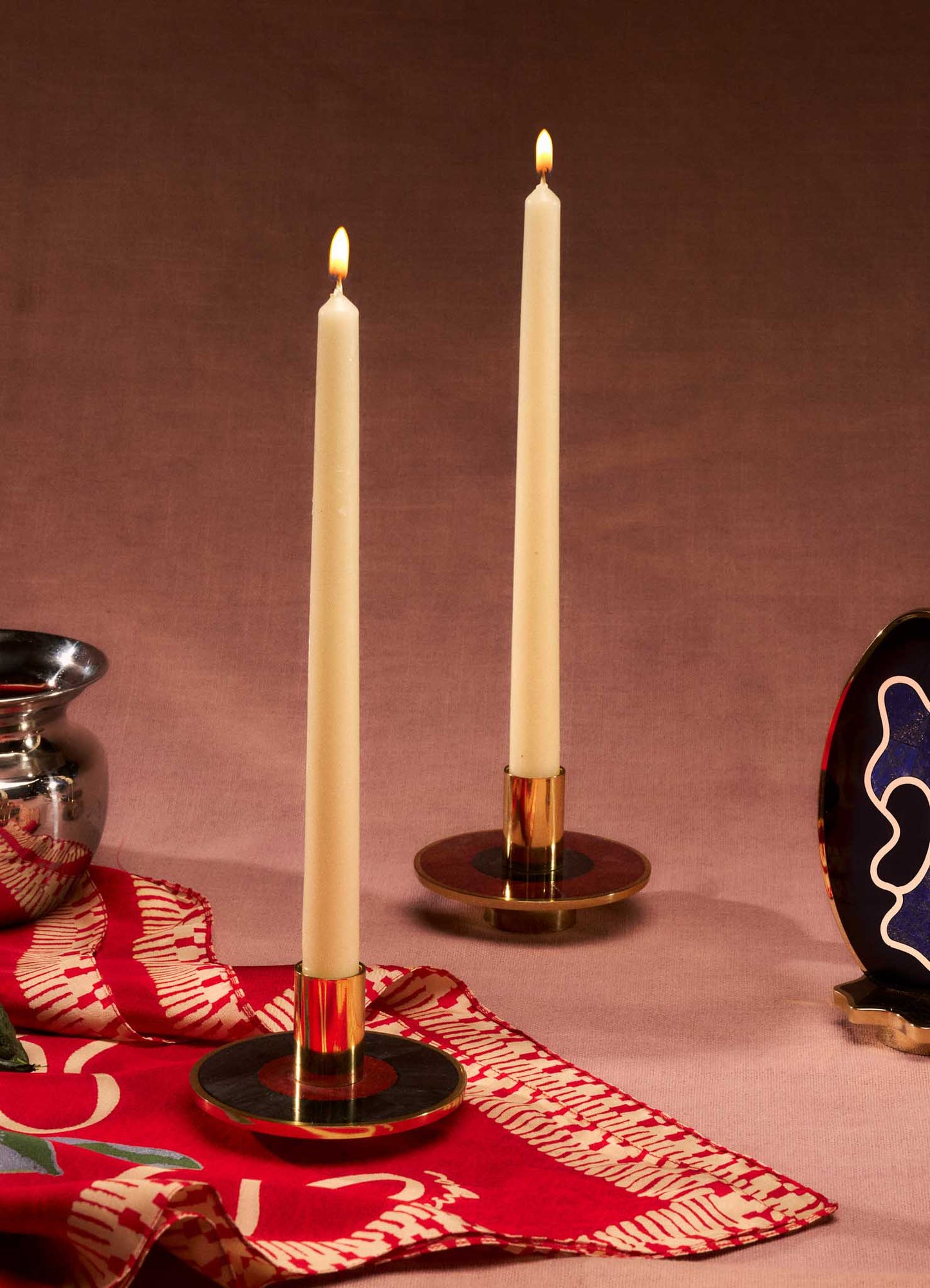 Brass candle holders with a circular base and polished gold finish, holding tall white taper candles with lit flames, set on a pink surface with a patterned red and white textile.