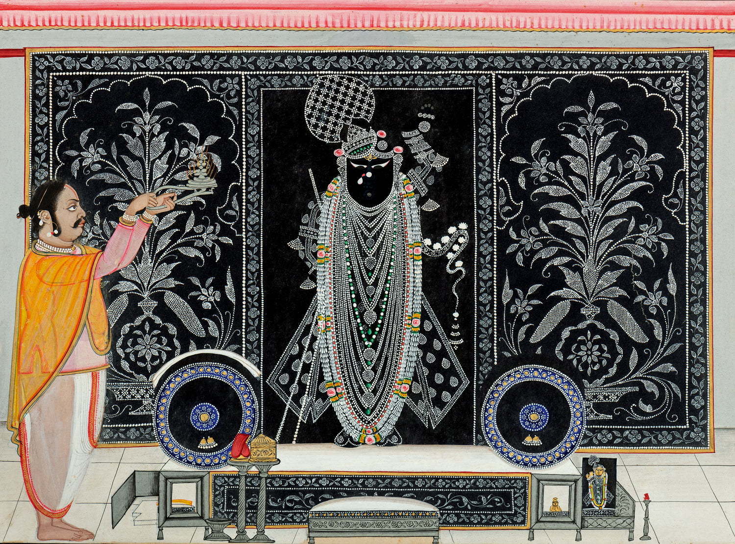 A traditional Indian wall hanging or tapestry featuring intricate embroidery and beadwork in black, white, and metallic accents. The design depicts a central deity figure adorned with jewelry, surrounded by floral motifs, with a person in traditional attire offering an offering in front.