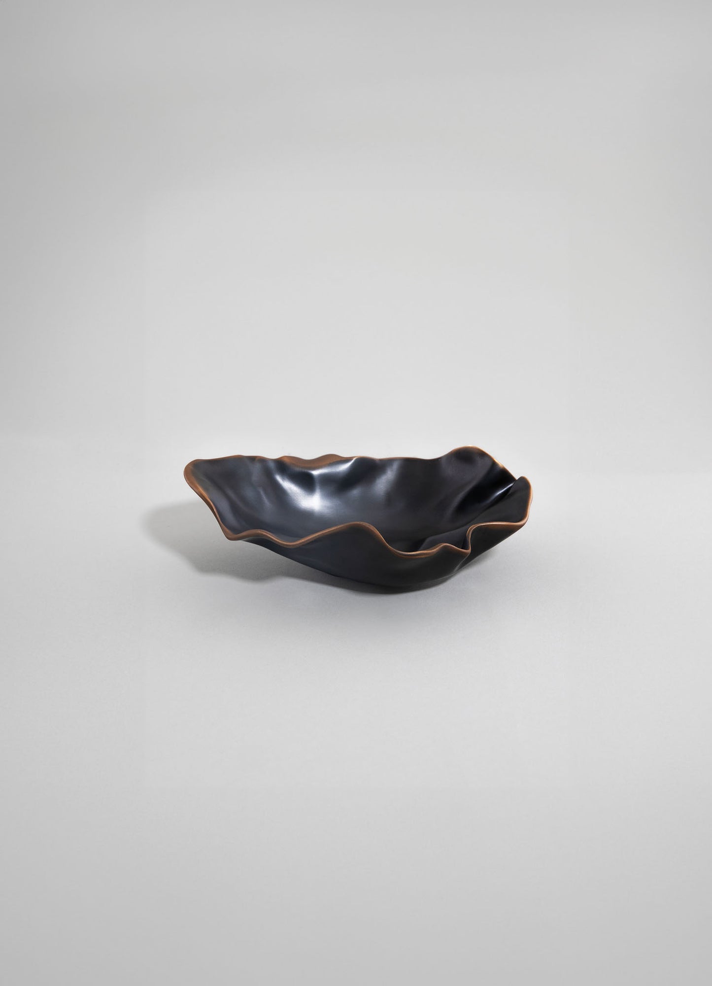 A ceramic dish with a wavy, ruffled edge and a glossy black finish, accented by a thin bronze-colored rim, set against a neutral background. The style is contemporary with an organic, sculptural form.