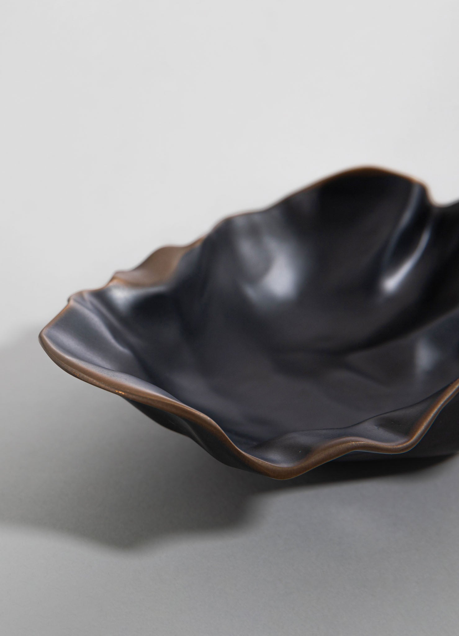 A black ceramic bowl with a ruffled, organic shape and a subtle brown edge detail, displayed on a neutral gray surface. The design emphasizes a handcrafted, contemporary style with a smooth, matte finish.